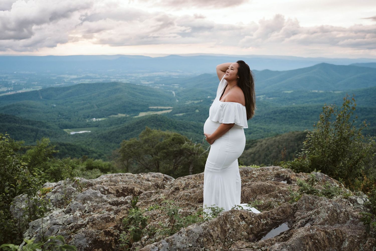 A serene mountain vista stretches into the distance as a figure in white stands on rocky outcrop during cloudy day.