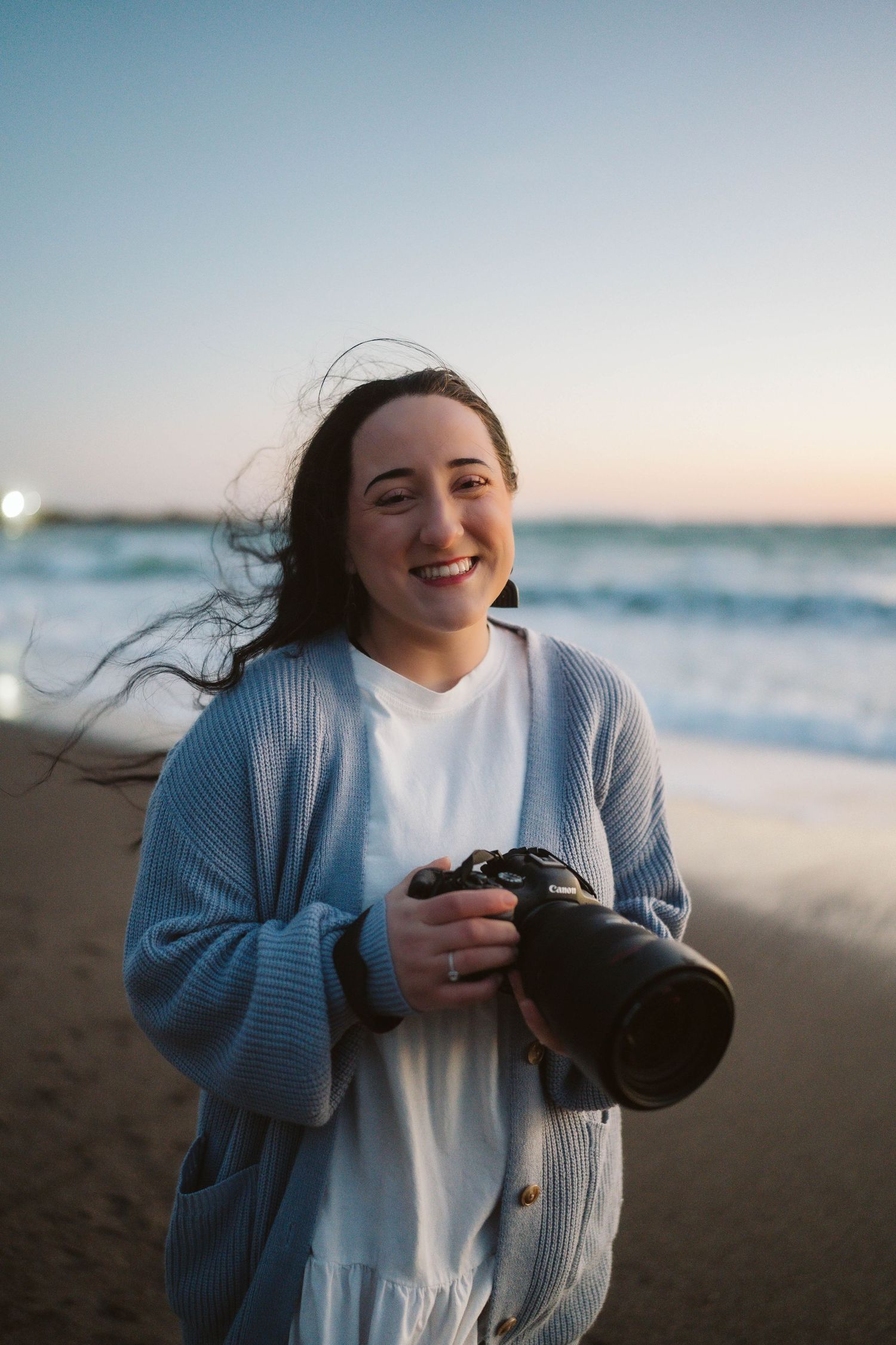 A photographer in a white dress and blue cardigan holds a camera on the beach during golden hour.