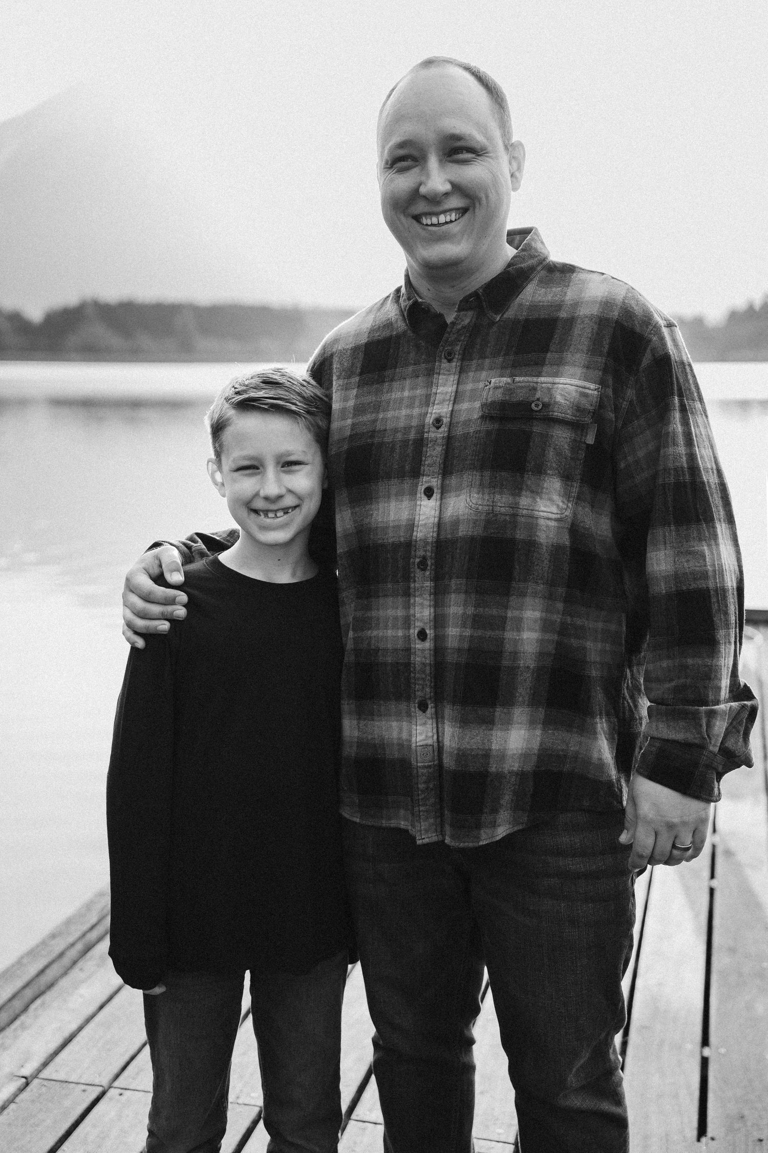 Black and white photo of a parent and child standing together on a wooden dock by a lake, wearing casual clothes.