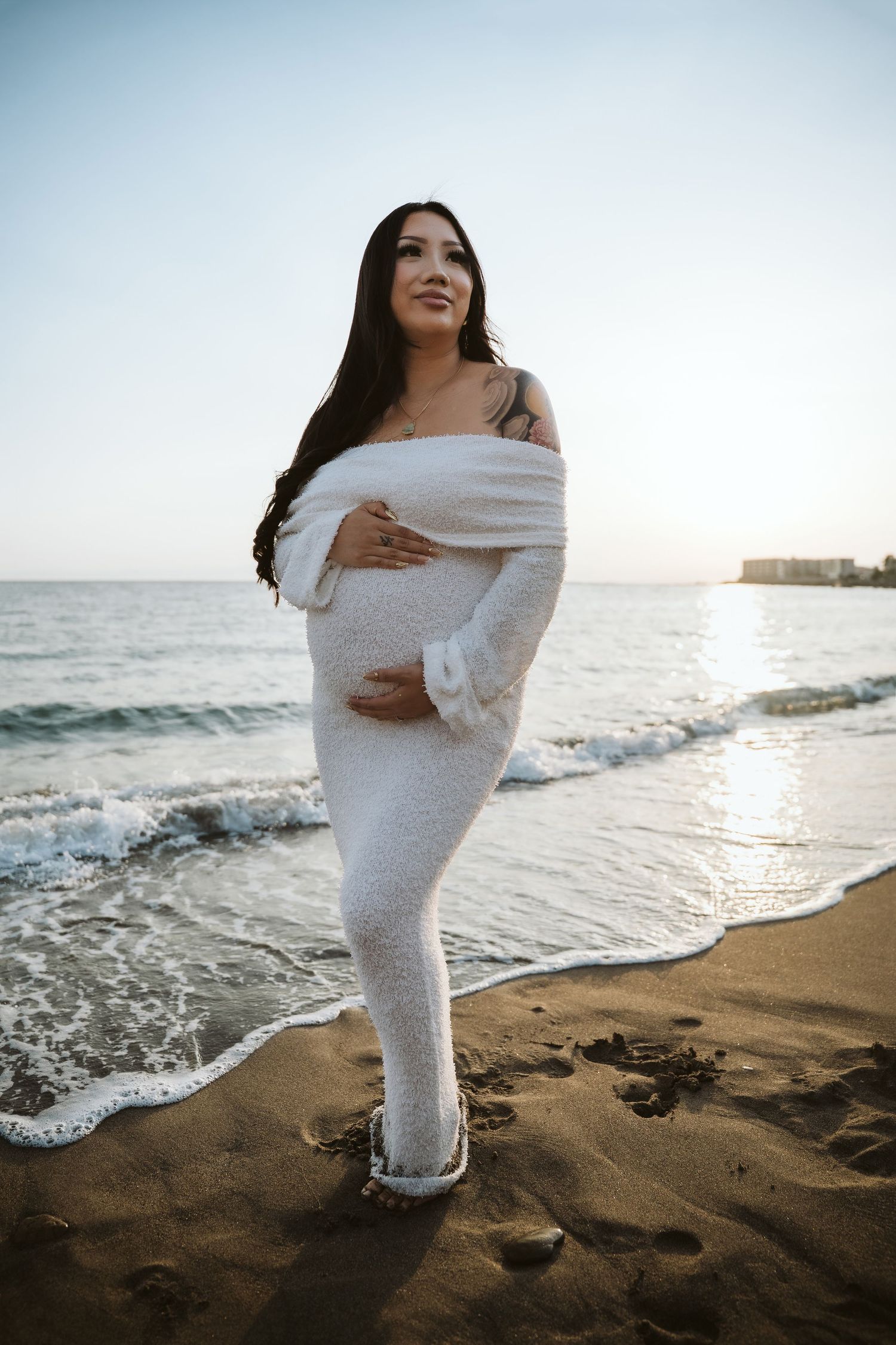 A pregnant woman in a white dress stands on a beach at sunset, looking towards the horizon.
