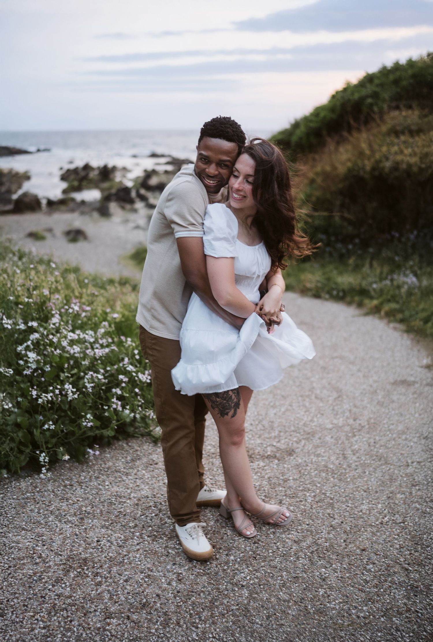 A couple shares a joyful embrace on a coastal path with rocky shores and wildflowers in the background at sunset.