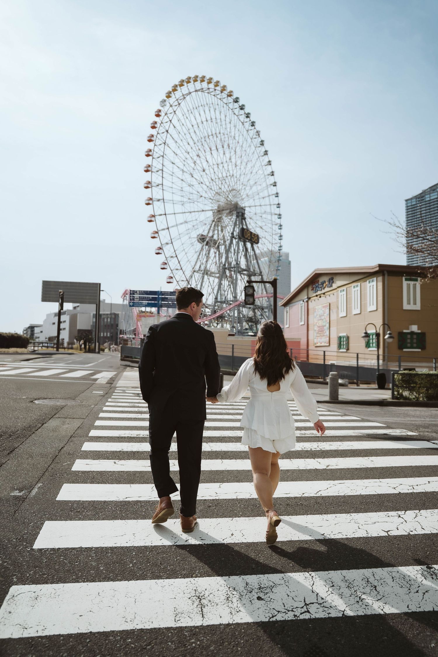 A couple walking across a crosswalk with a large ferris wheel visible in the background.