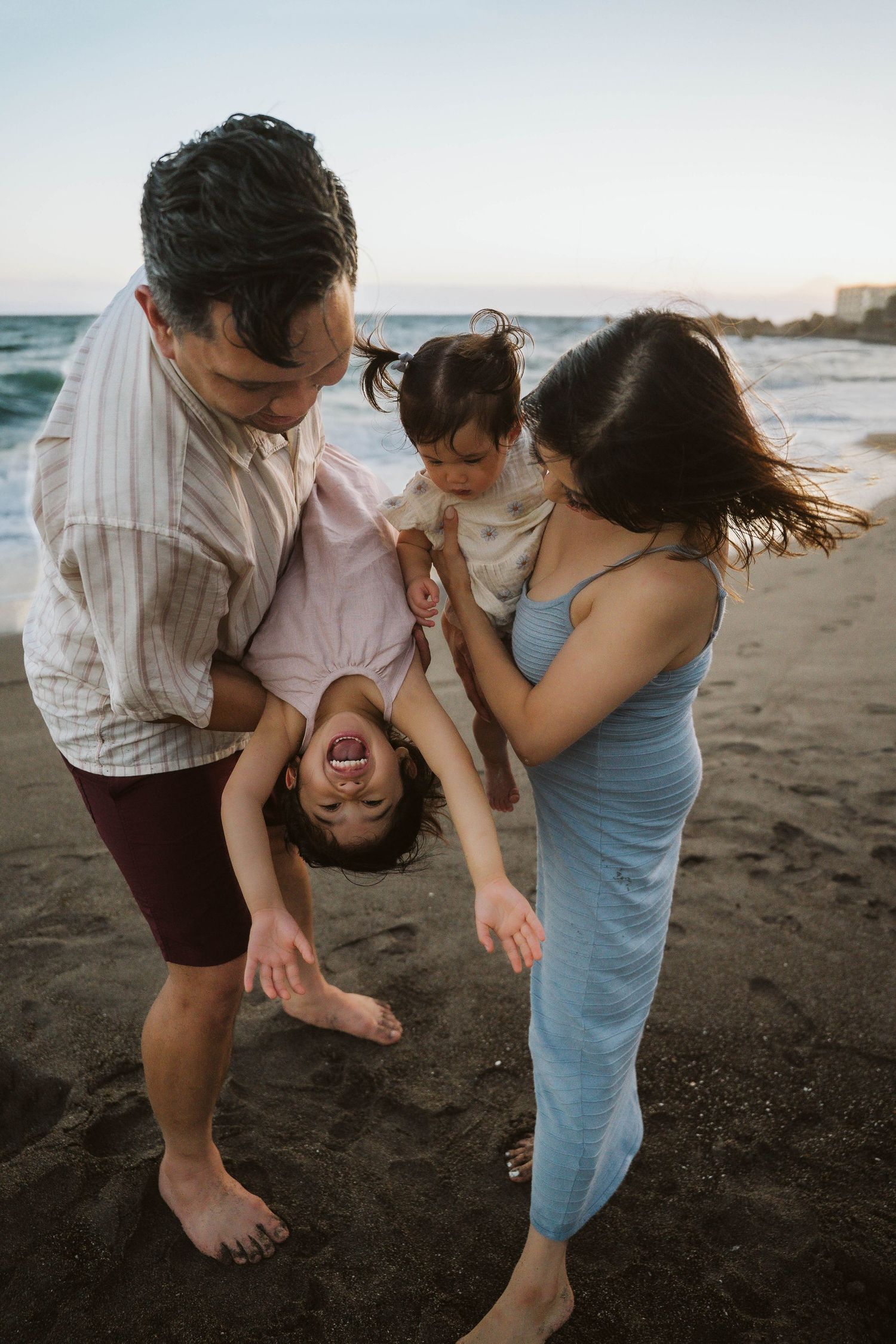 A loving family plays together on a dark sandy beach at sunset with waves crashing in the background.