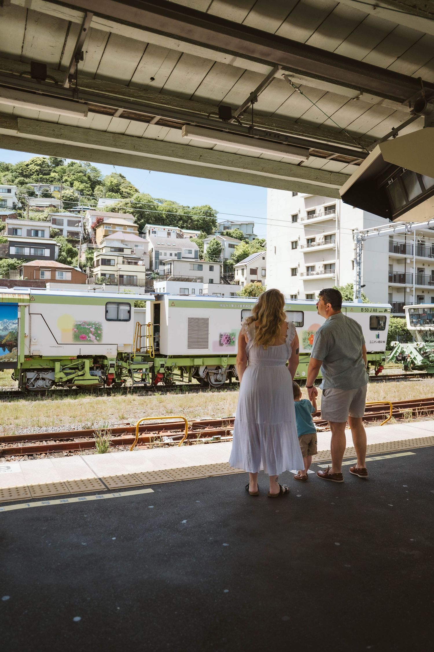 People wait on a covered train platform overlooking residential buildings on a hillside.