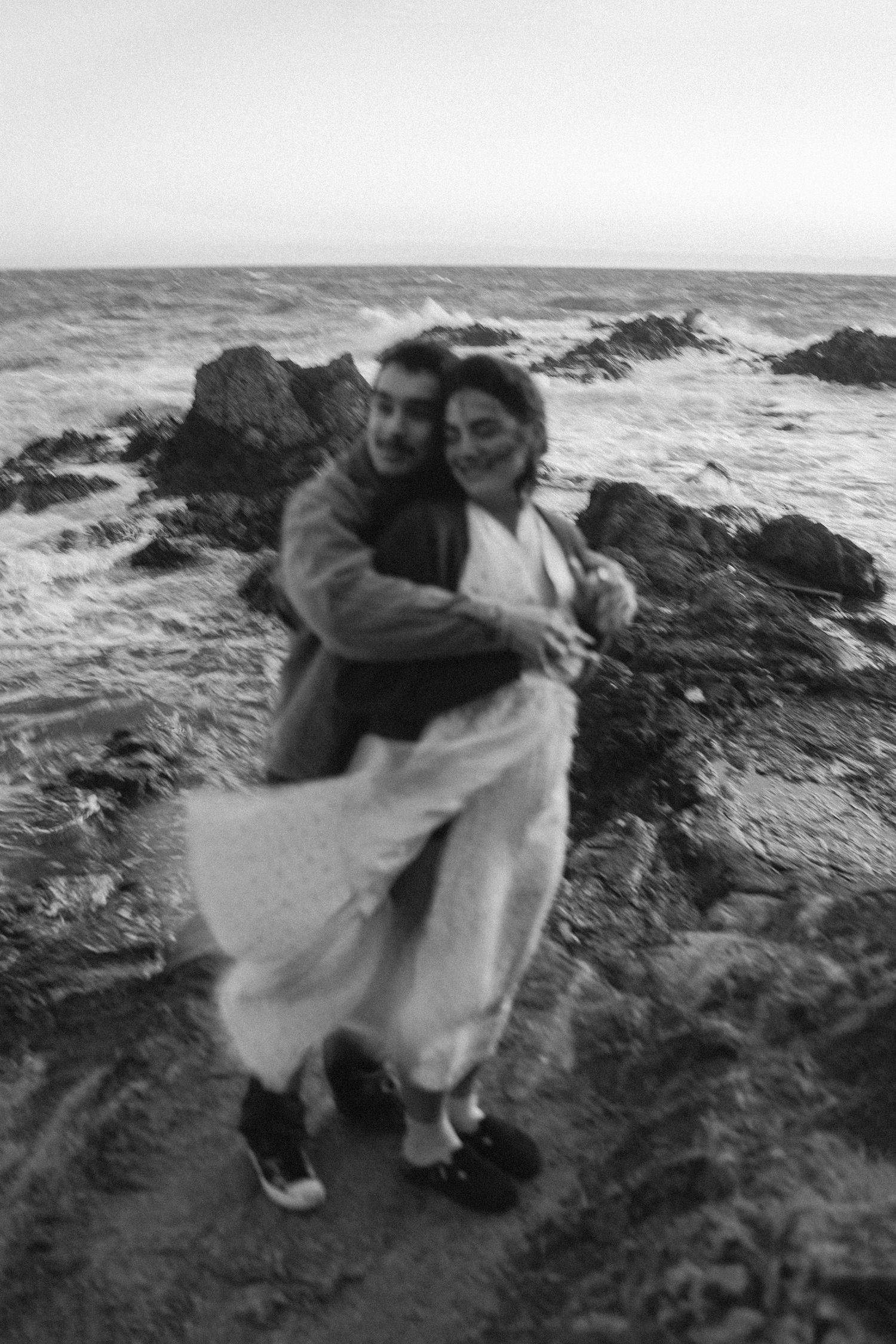 A romantic black and white photo of a couple embracing on rocky coastal cliffs by the sea.