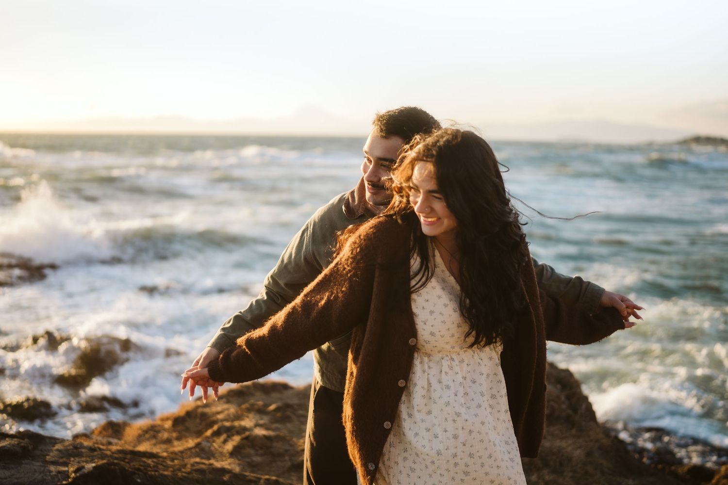 Couple shares joyful moment on rocky coastline with crashing waves and golden sunlight.