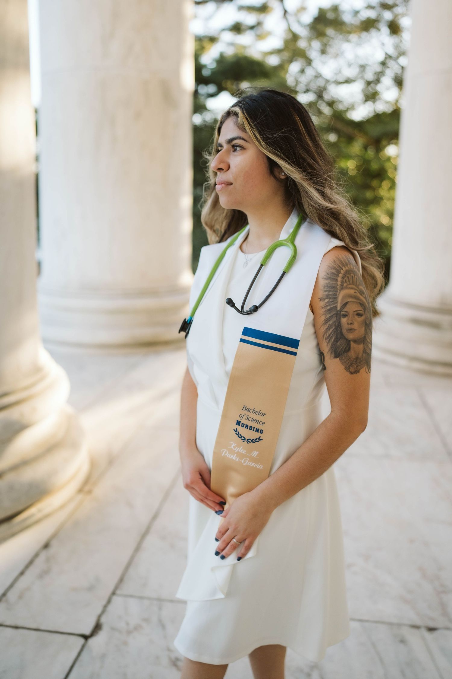 Healthcare professional in white dress with stethoscope standing by classical columns in outdoor setting.