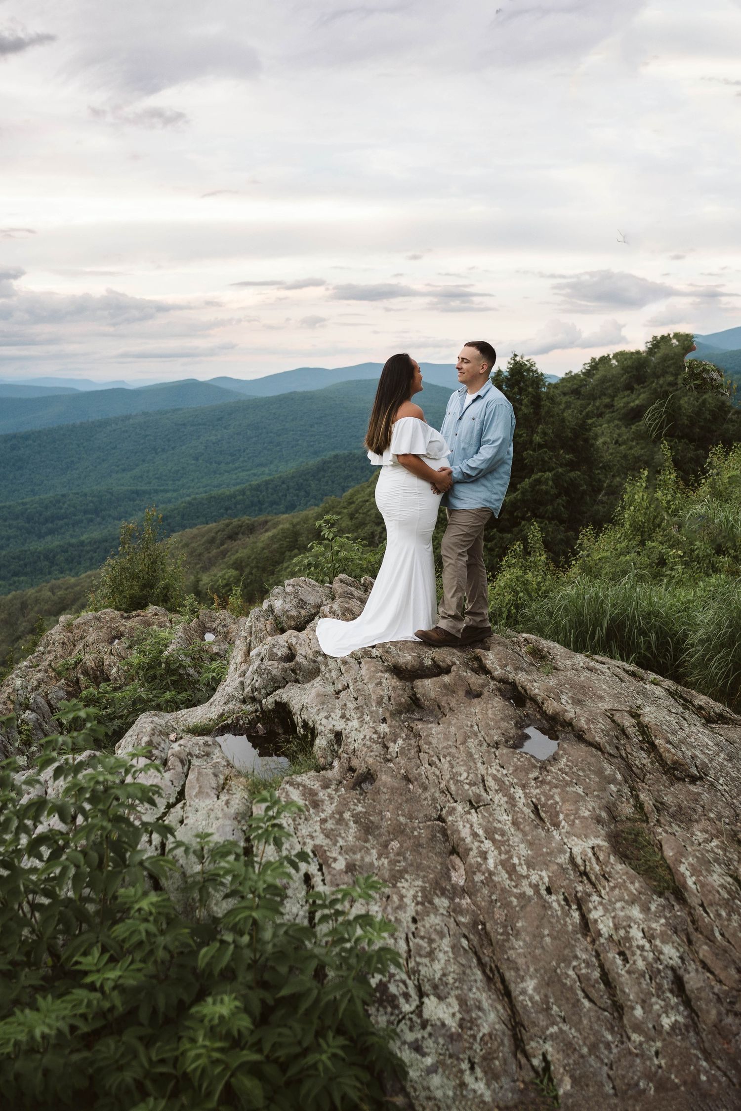 A couple stands together on a rocky mountain overlook with rolling blue ridges and cloudy skies in the background.
