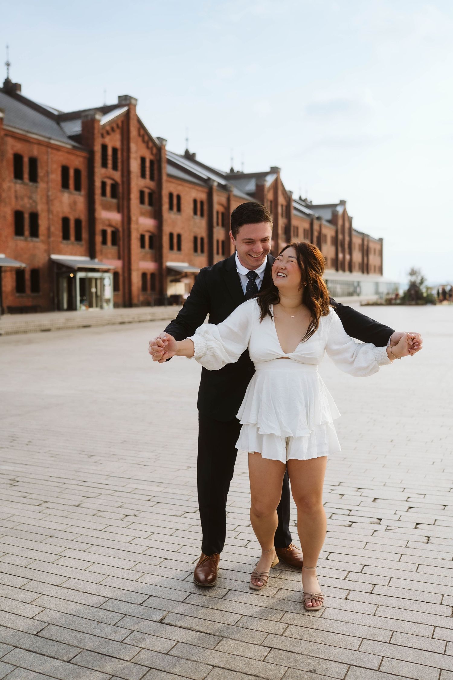 A loving couple embraces and laughs together on a brick-lined path outside historic red brick warehouses.