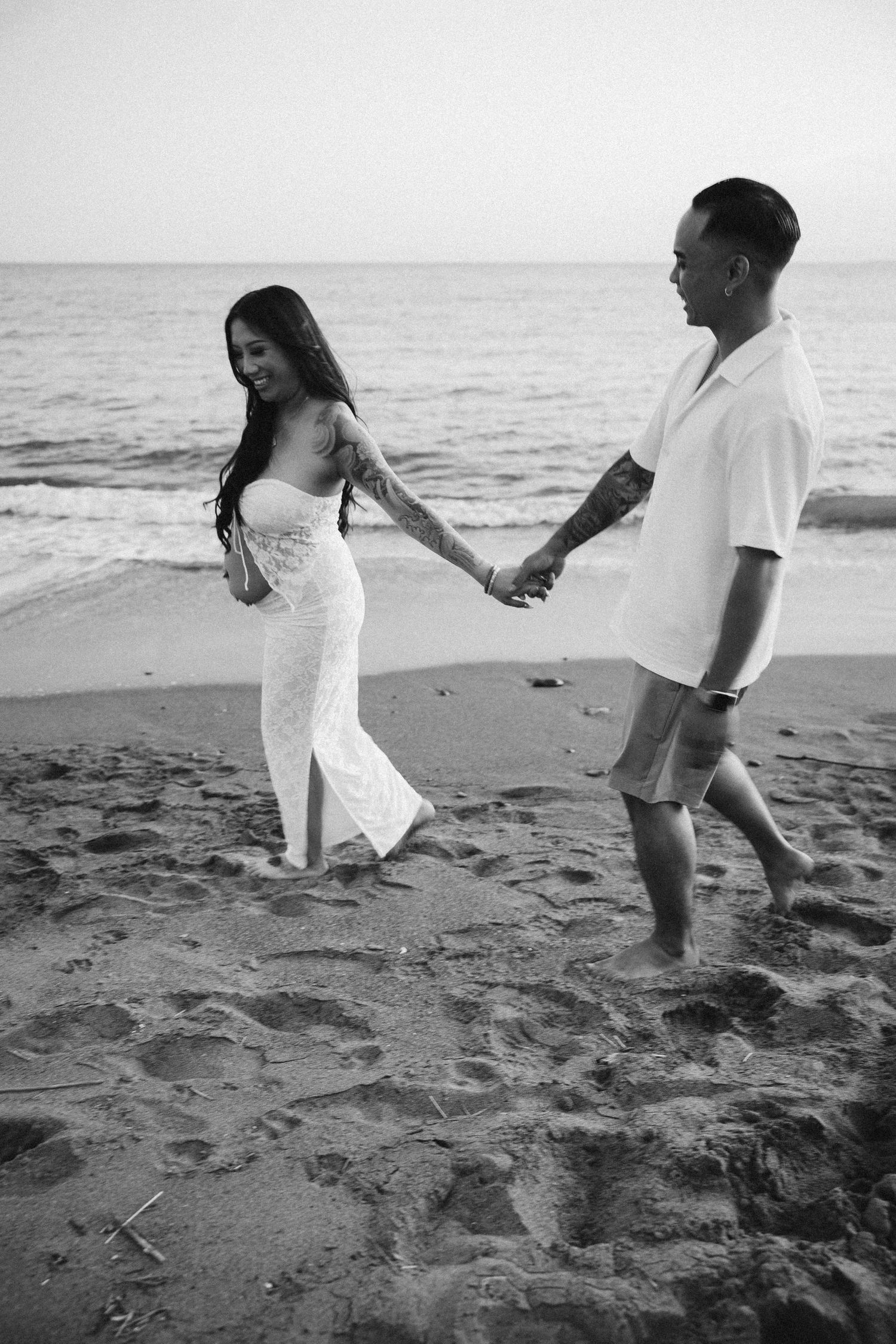 A romantic black and white photo of a couple walking hand in hand on a sandy beach.
