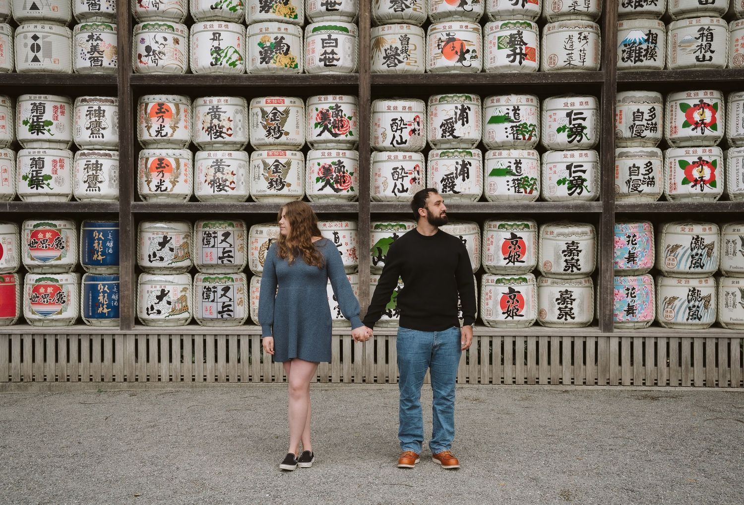 Rows of traditional Japanese sake barrels with decorative designs line a wall while visitors view the display.