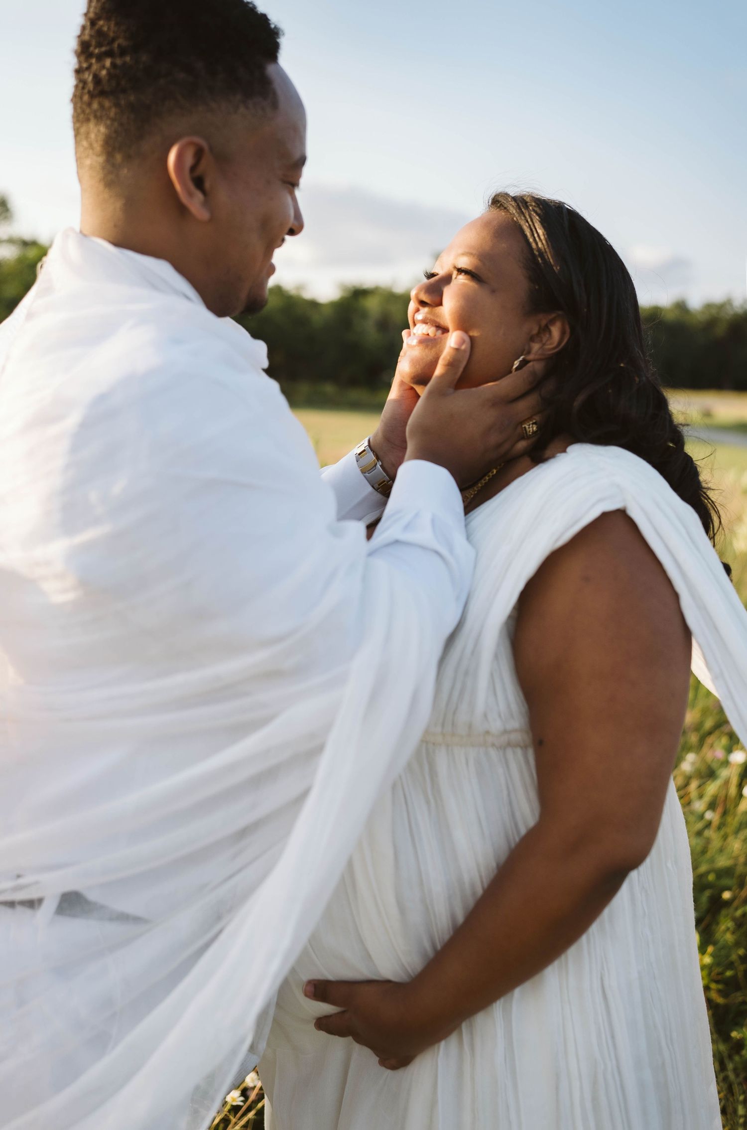 A tender moment between a couple in white attire embracing in a sunlit field during a wedding photoshoot.