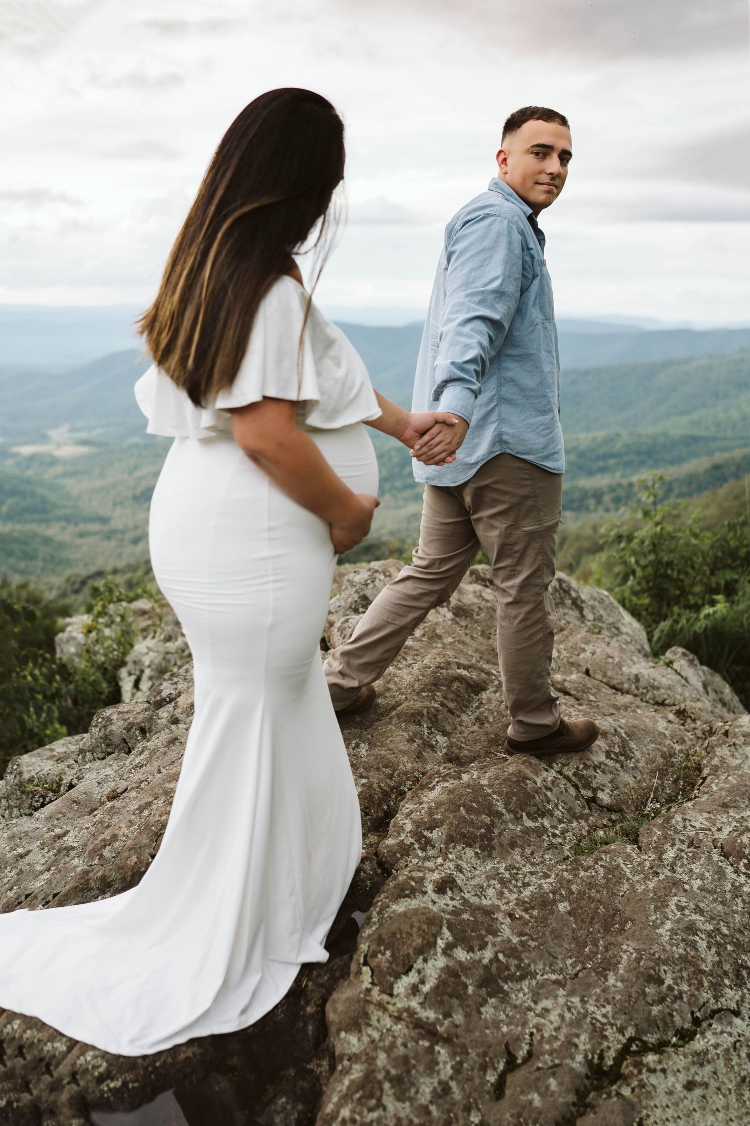 A couple stands on a mountain peak overlooking a scenic valley, with the woman wearing a flowing white dress.