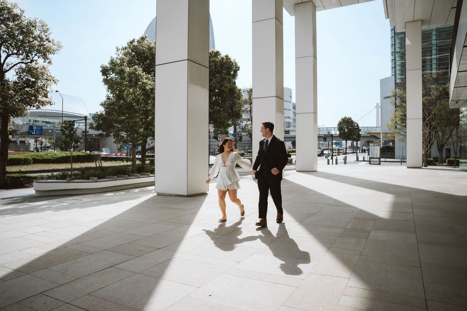 A couple walks through modern white pillars and archways of a sleek building while casting shadows on the tiled floor.
