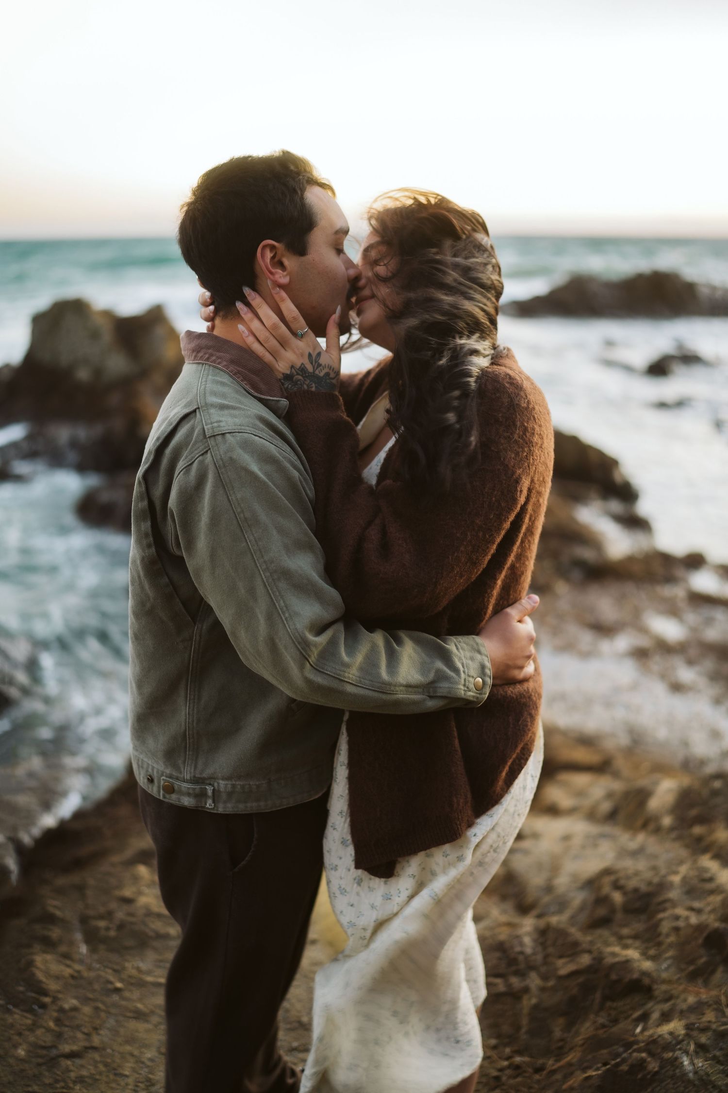 Romantic coastal photoshoot with couple embracing against dramatic ocean backdrop.
