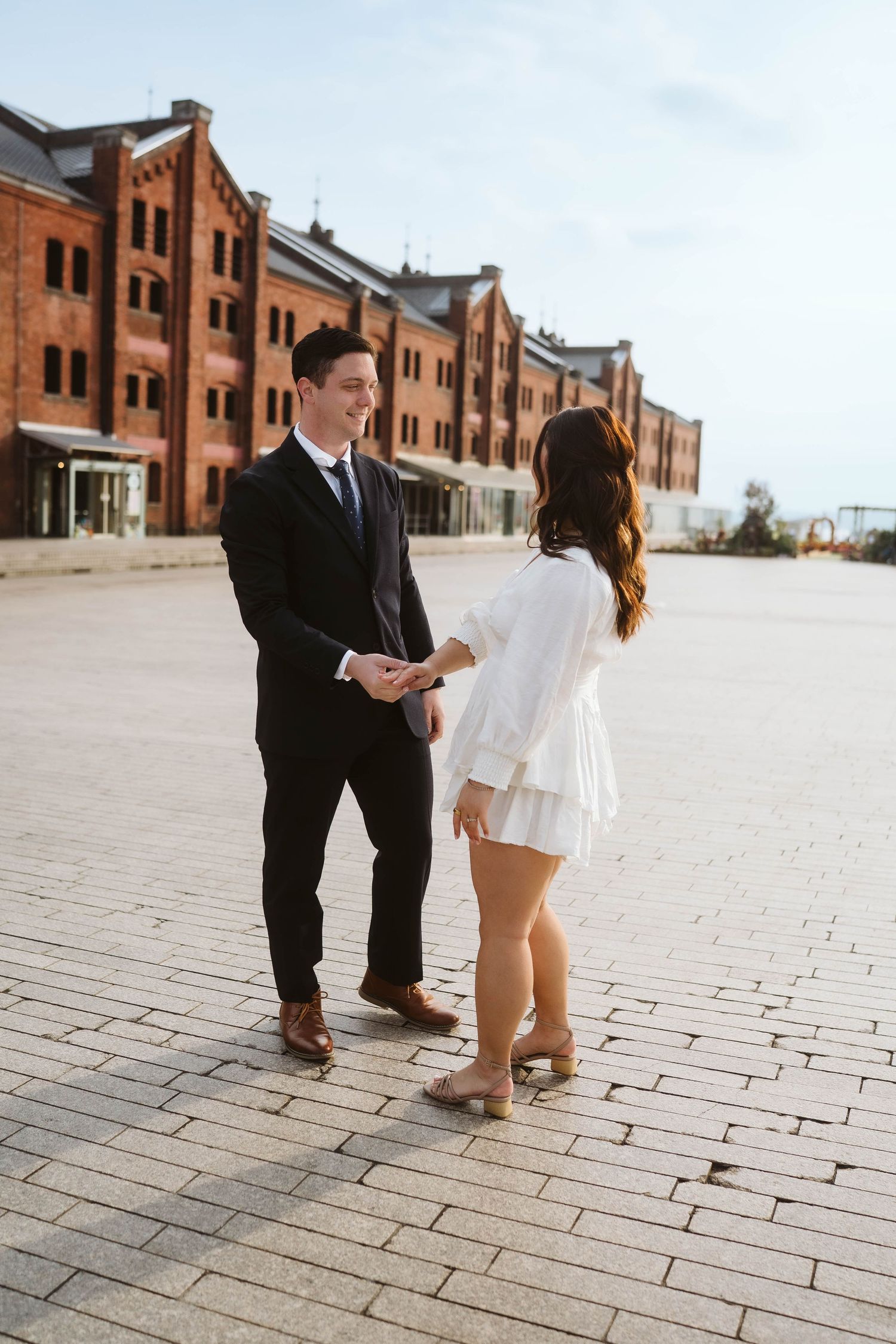 A romantic couple stands together on brick pavement in front of historic red brick warehouses on a sunny day.