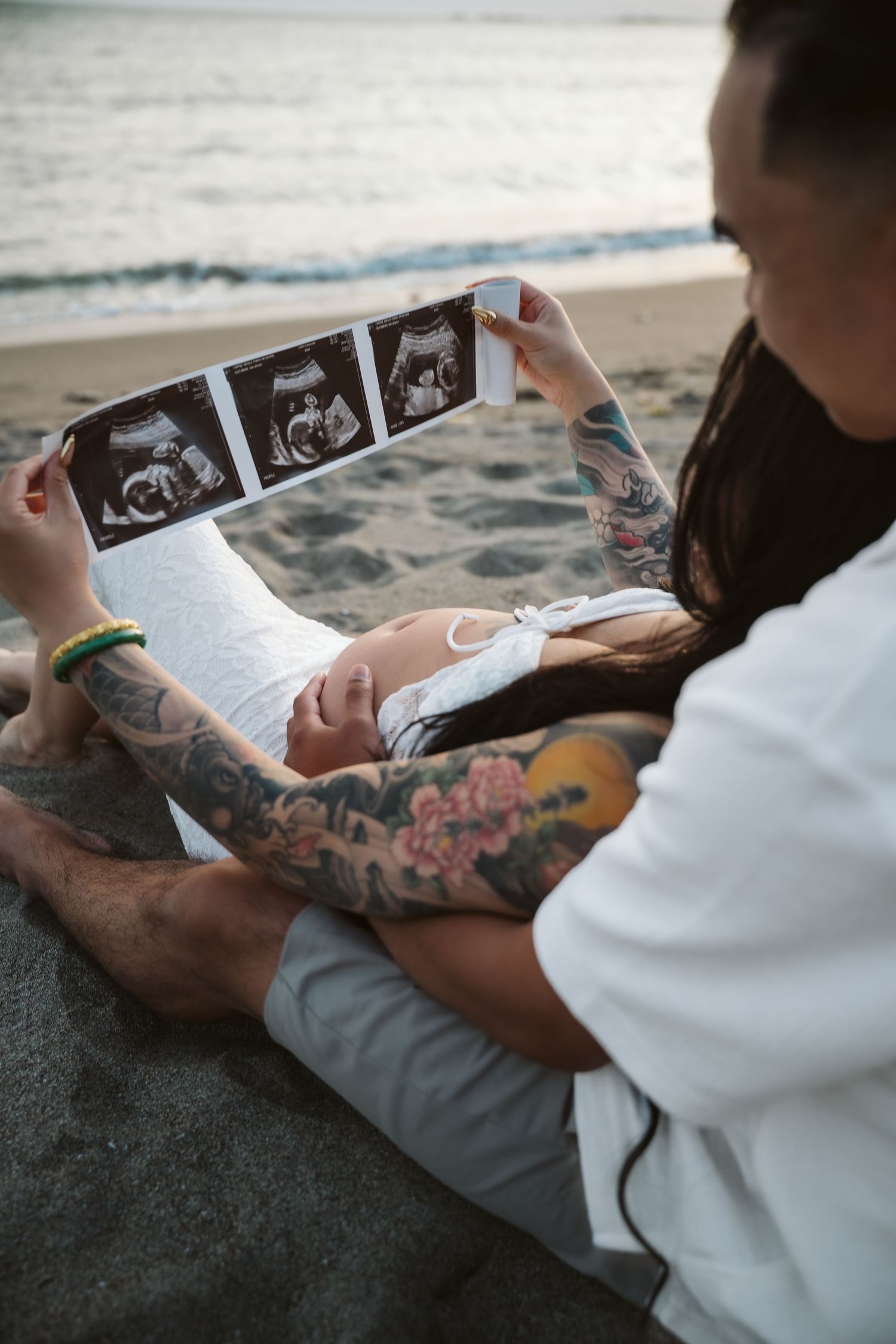 A couple looking at ultrasound photos while relaxing on a beach at sunset, with visible arm tattoos.
