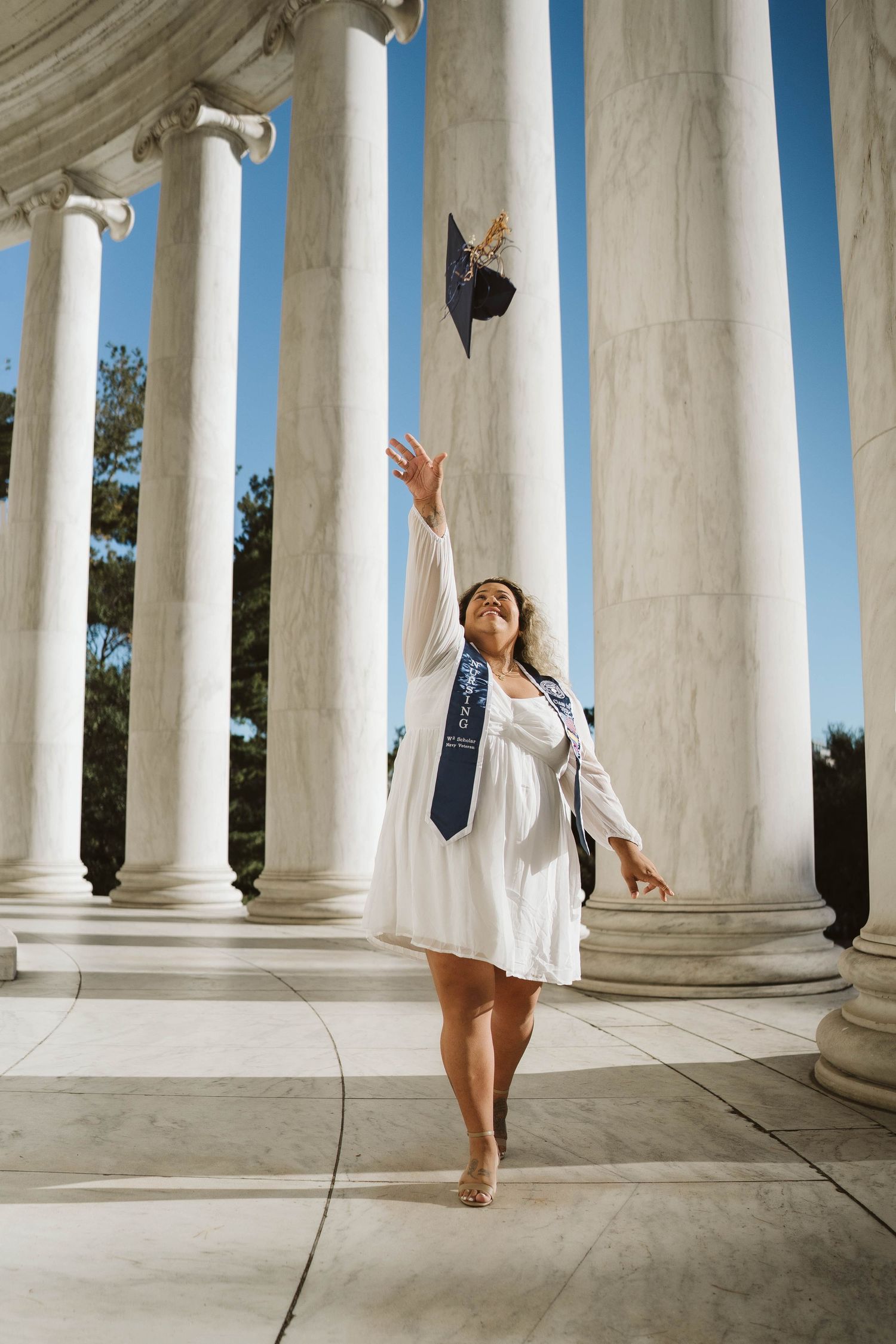 Graduate in white dress throws graduation cap in the air while standing between classical white marble columns.