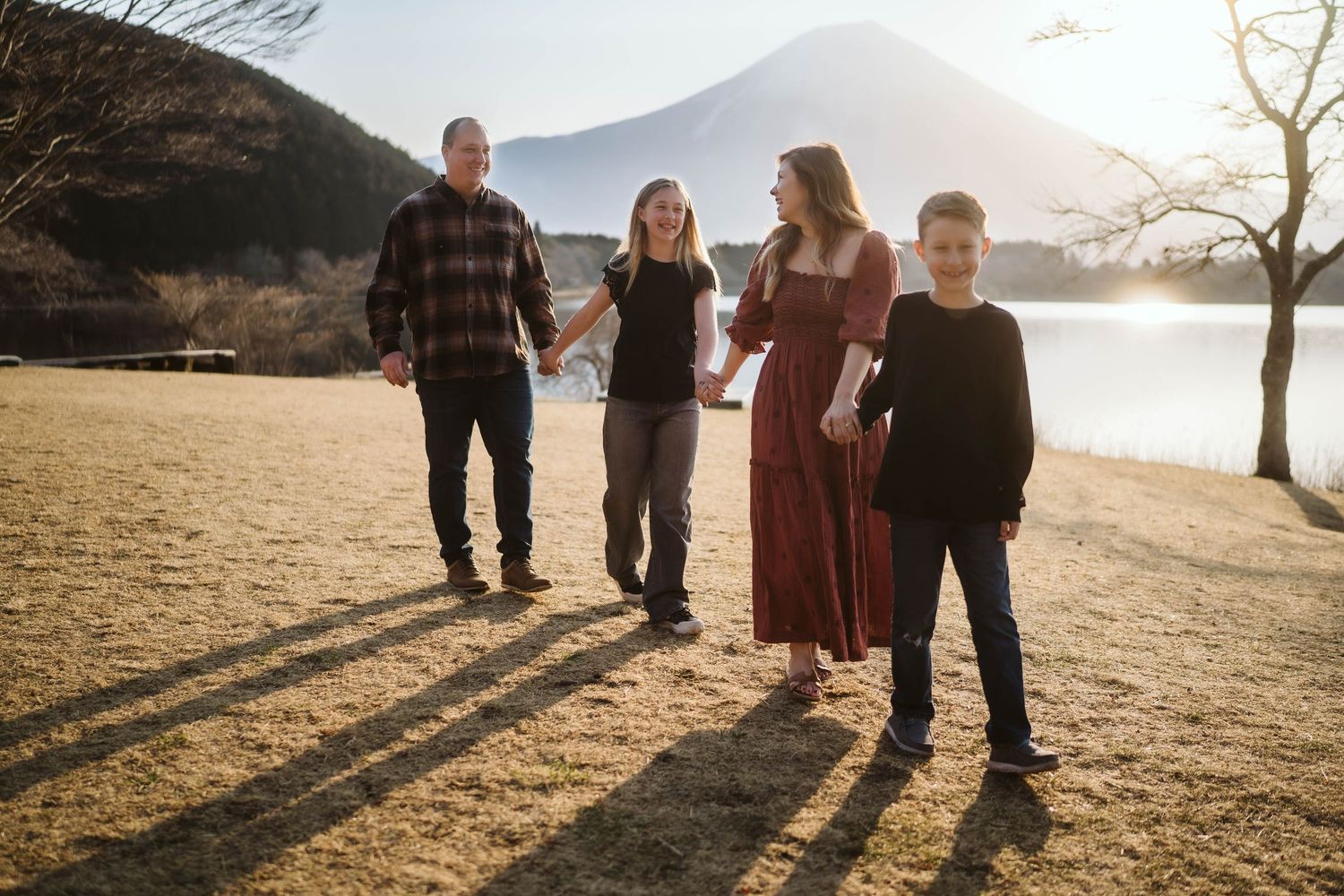 A family stands together on grassy terrain near a lake at sunset casting long shadows on the ground.