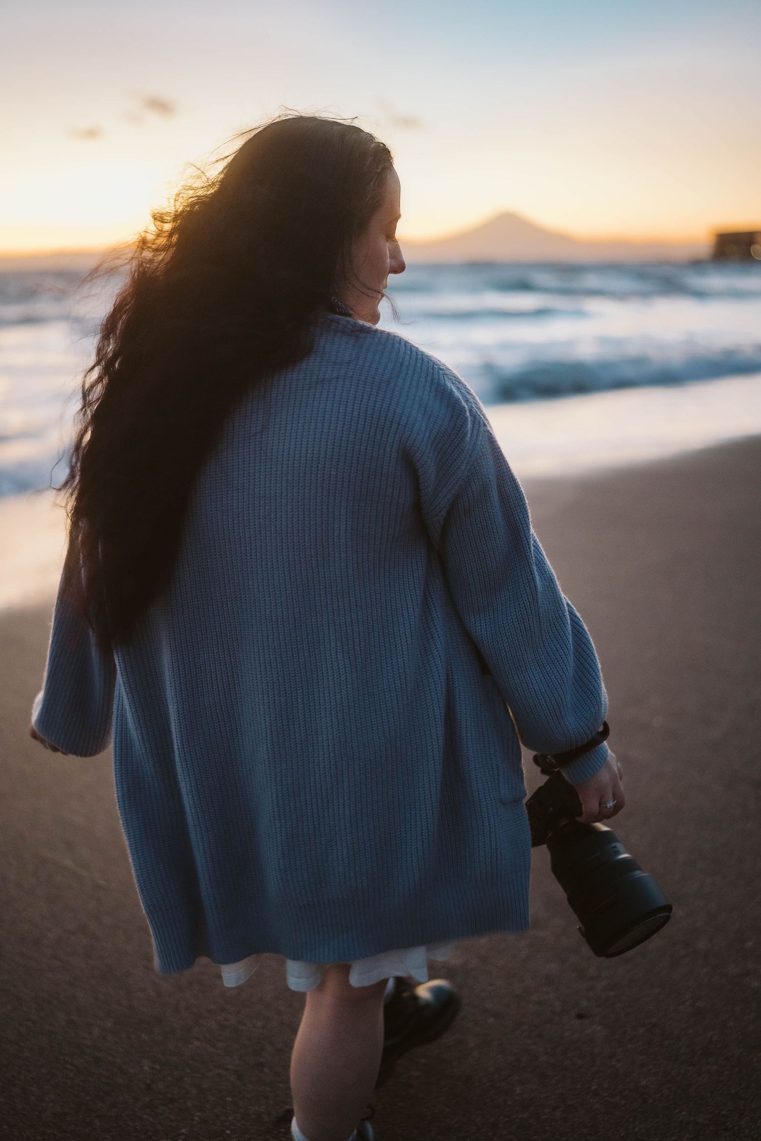 Silhouetted figure in blue sweater walks on beach at sunset with waves and mountain in background.