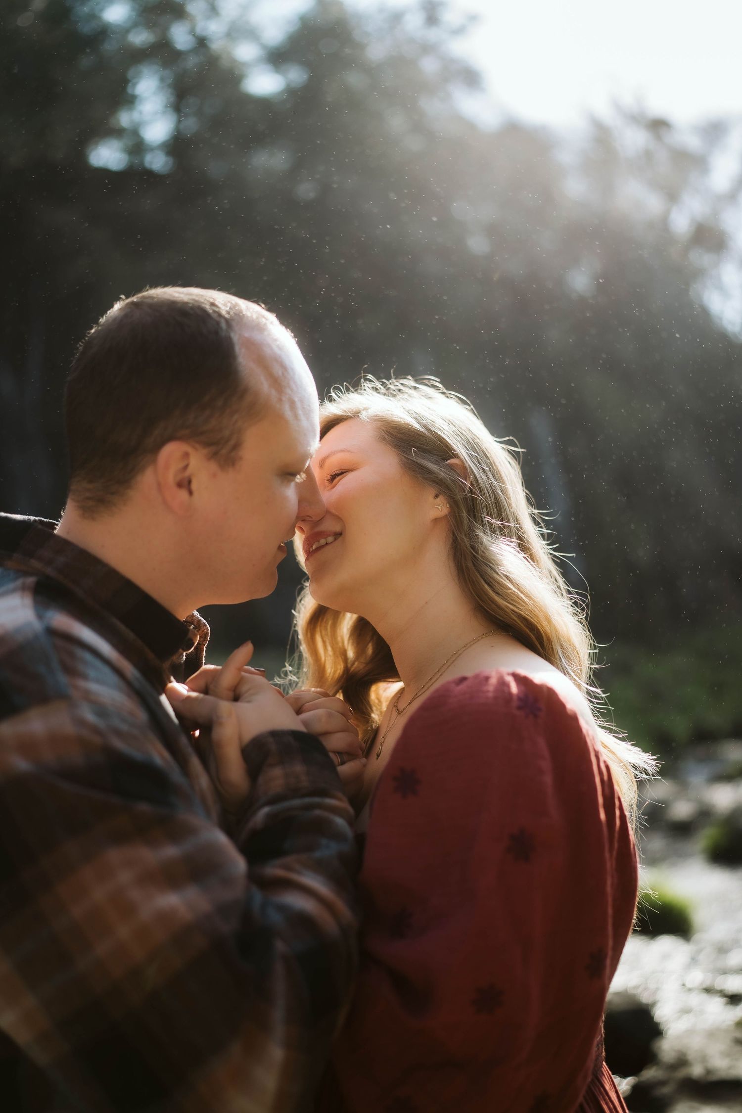 A romantic couple embraces in soft sunlight with trees and nature in the background.