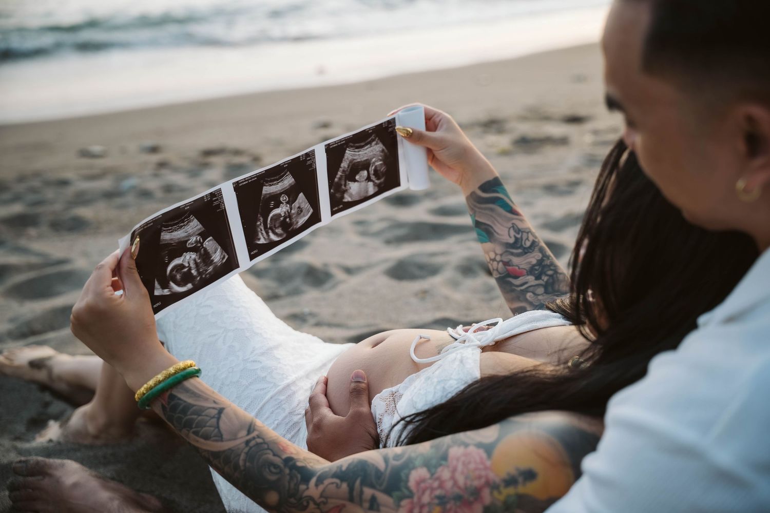 A couple sitting on the beach looking at ultrasound photos while sharing an intimate moment together.