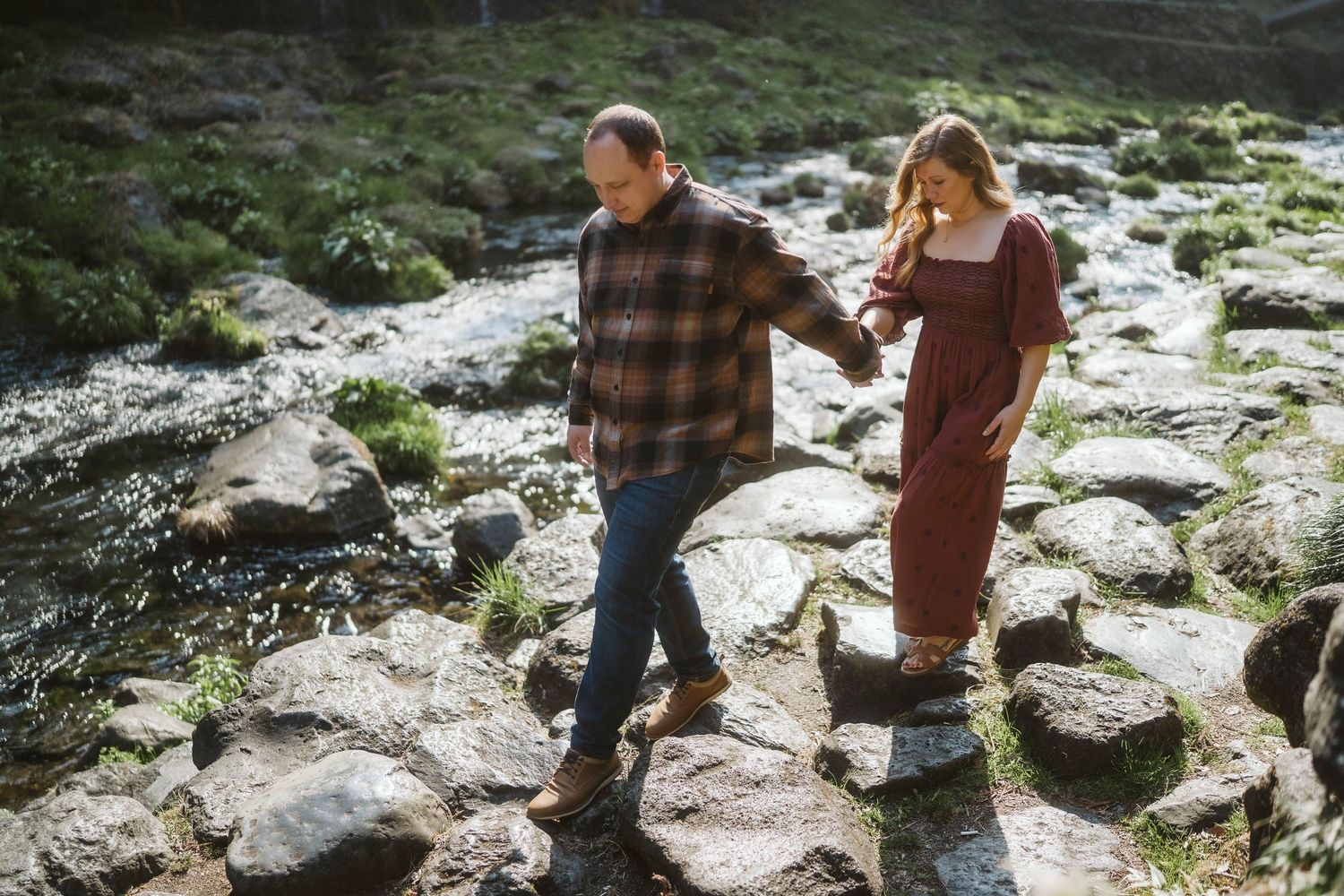 Two people walk hand in hand across rocky terrain on a sunny day, one wearing plaid and one in a rust-colored dress.