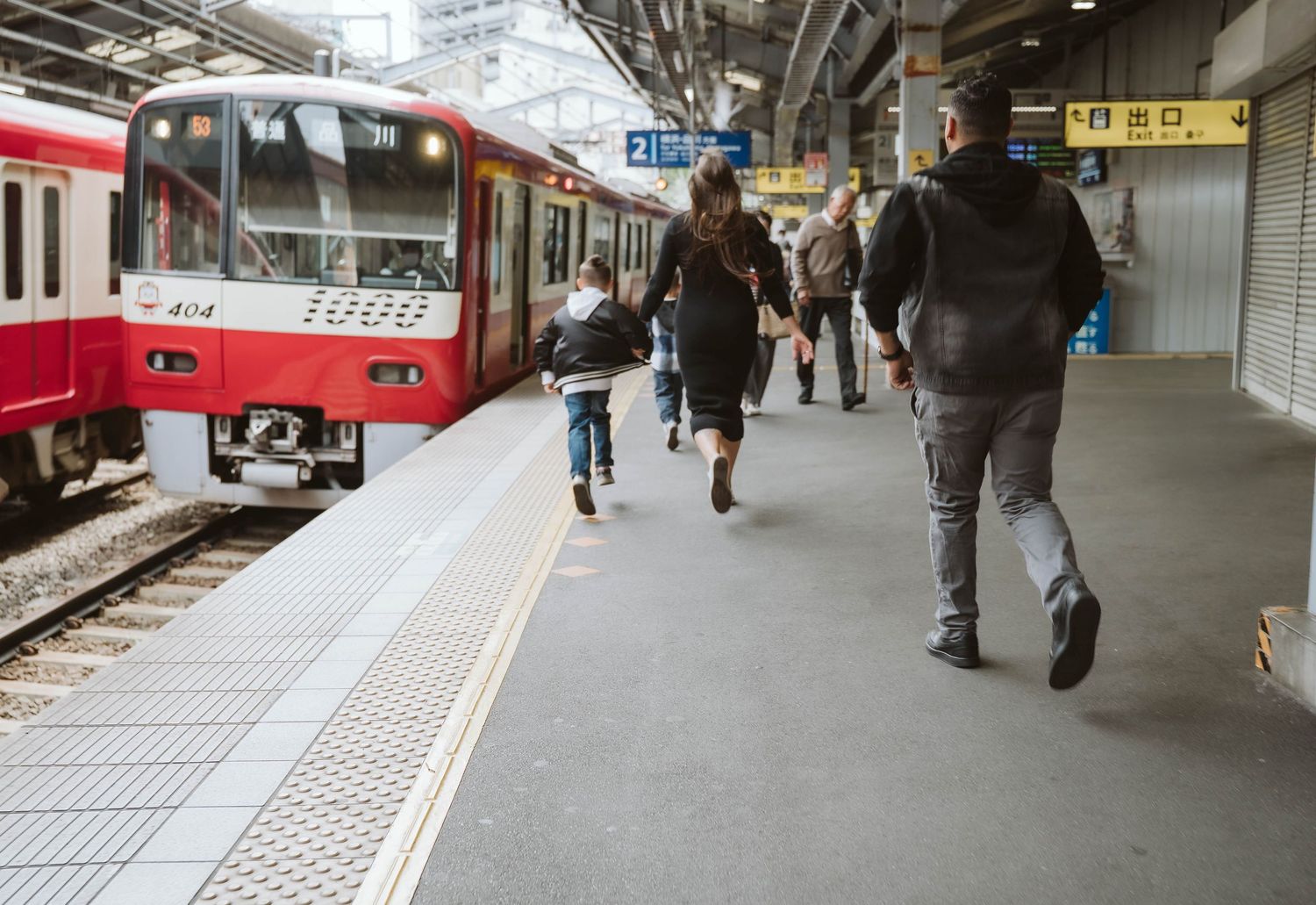 Passengers walk along a train platform next to a red commuter train in a Japanese railway station.