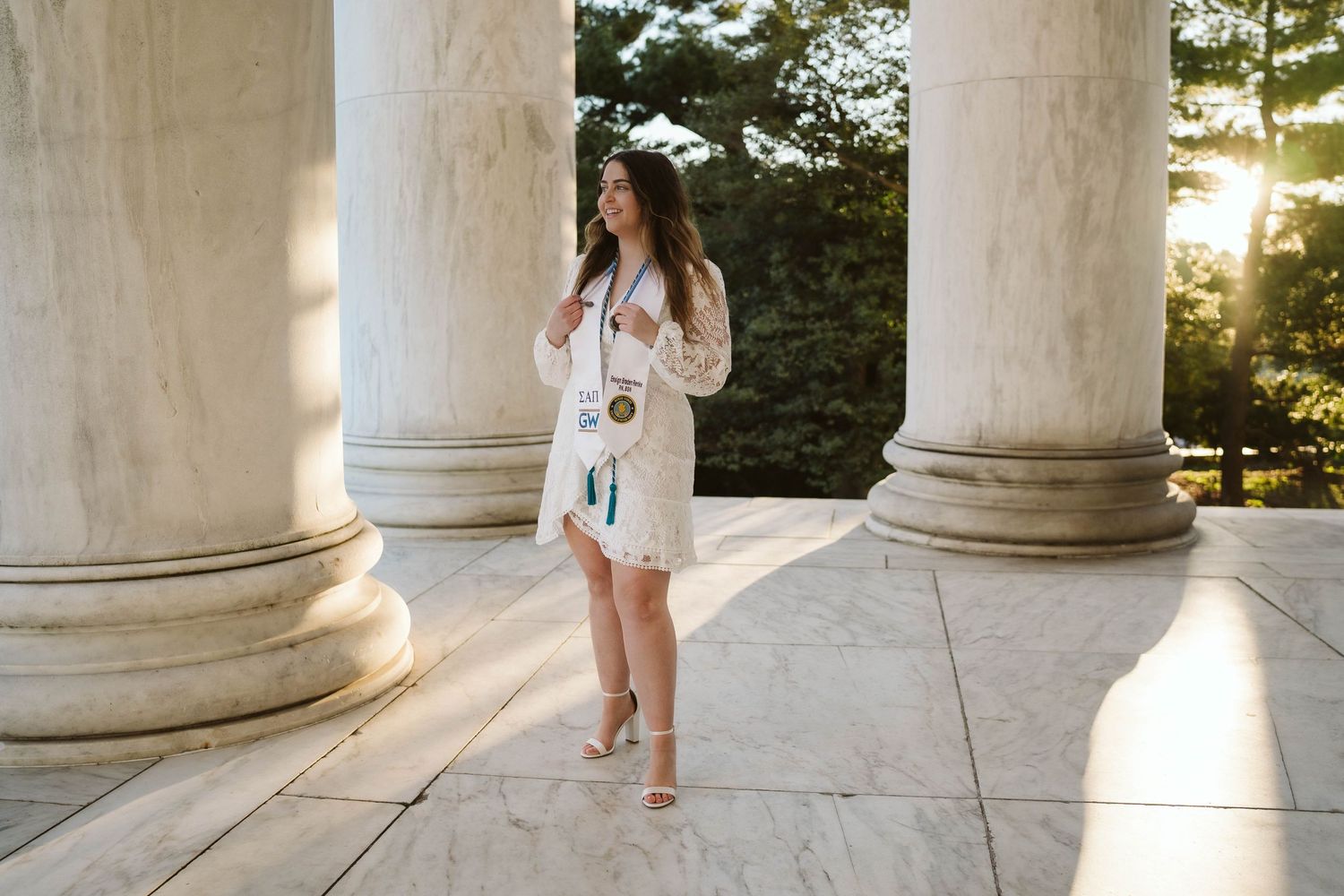 Someone in a white floral dress stands barefoot between classical columns as sunlight streams through at golden hour.