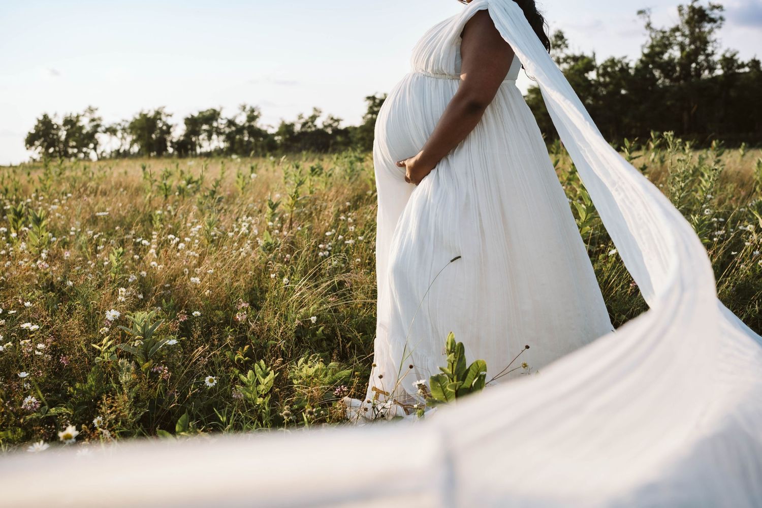 White flowing maternity dress billows in the wind as someone stands in a wildflower meadow during golden hour.