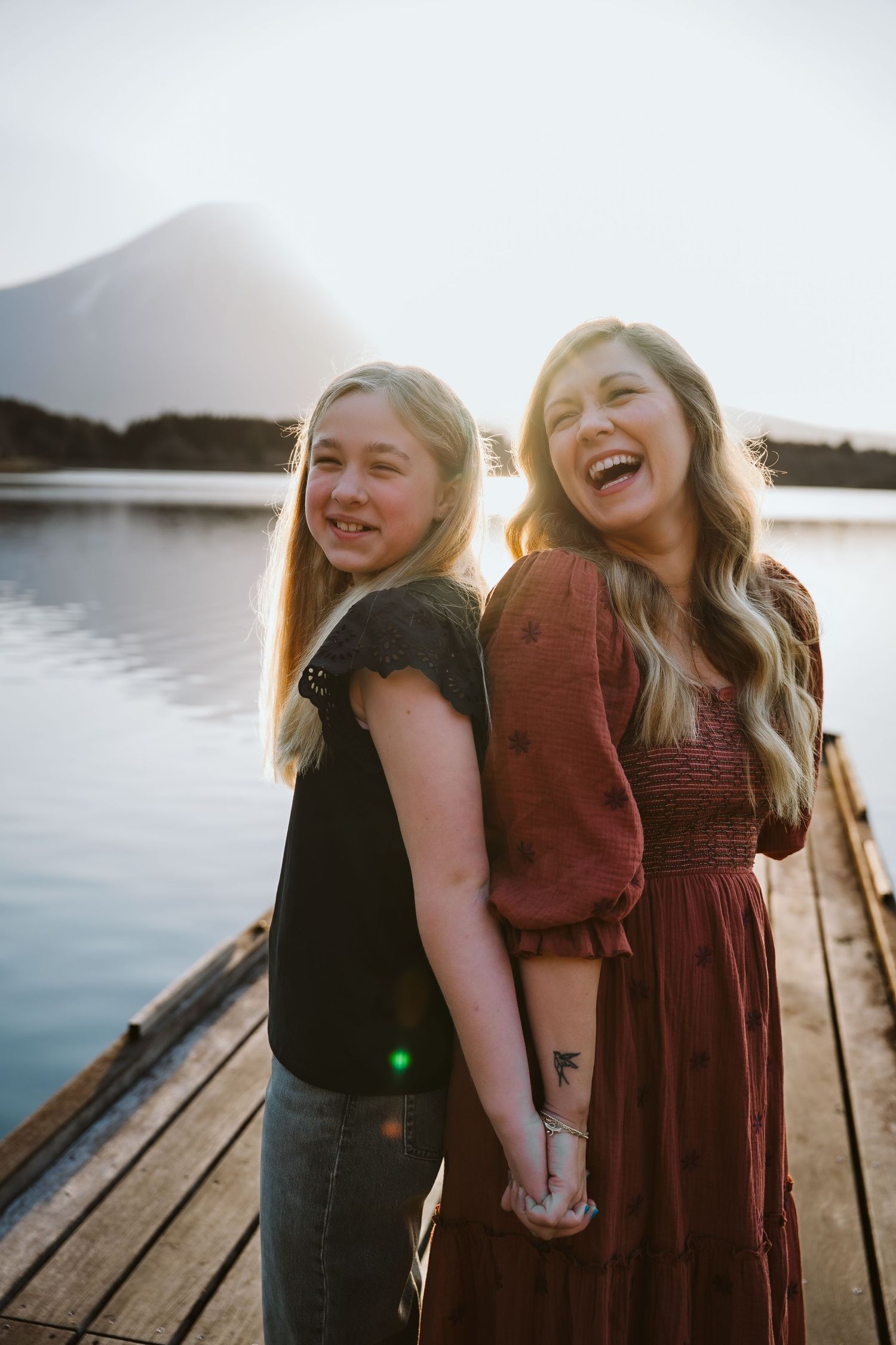 Two friends laugh together during sunset on a wooden dock by a mountain lake.