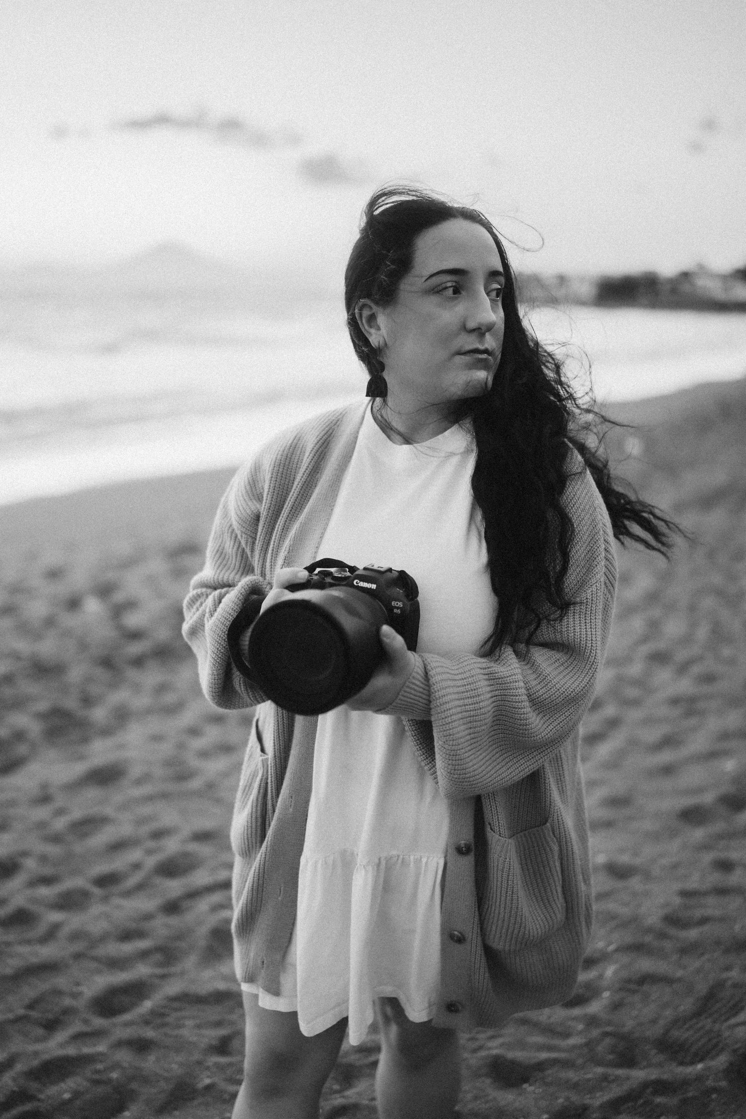 A contemplative photographer wearing a white dress and beige cardigan stands on a beach at sunset holding a camera.