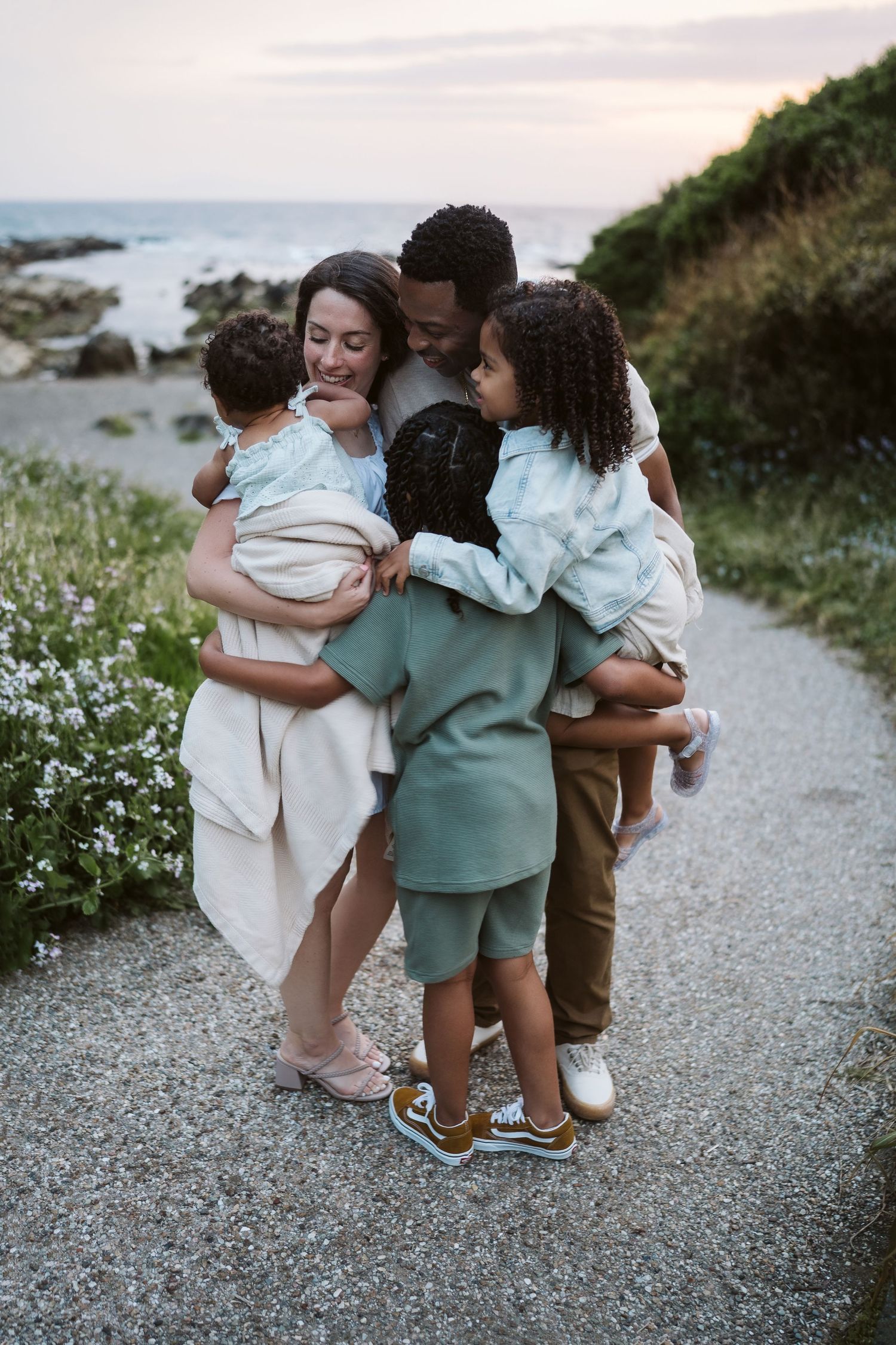 Family shares tender moments together on a coastal path surrounded by wildflowers at sunset.