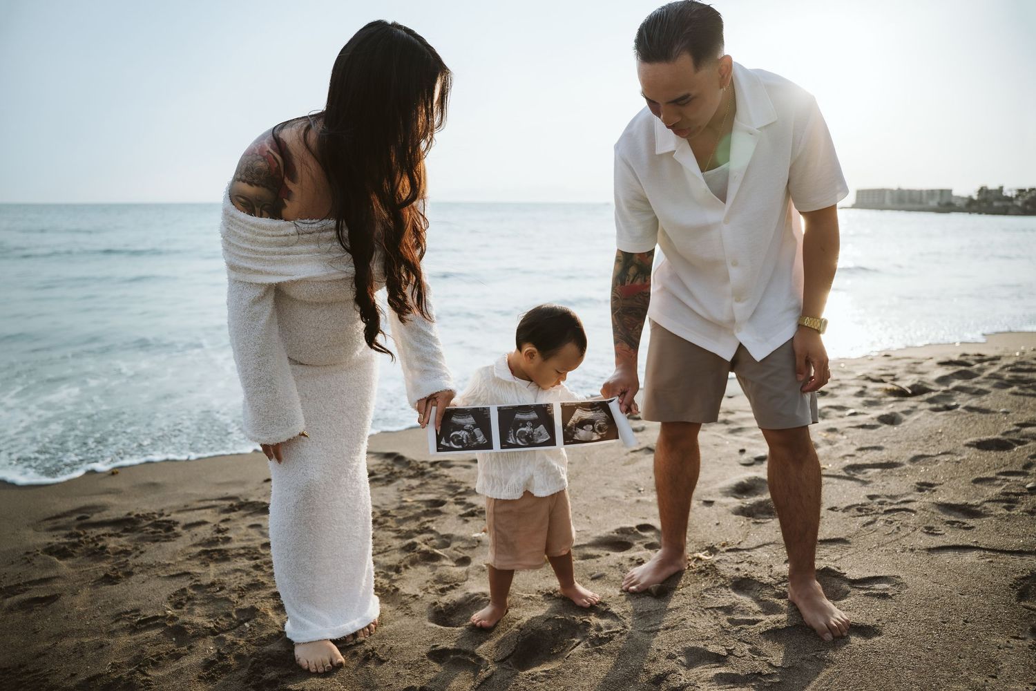 A family stands together on a beach holding ultrasound photos while making a pregnancy announcement at sunset.