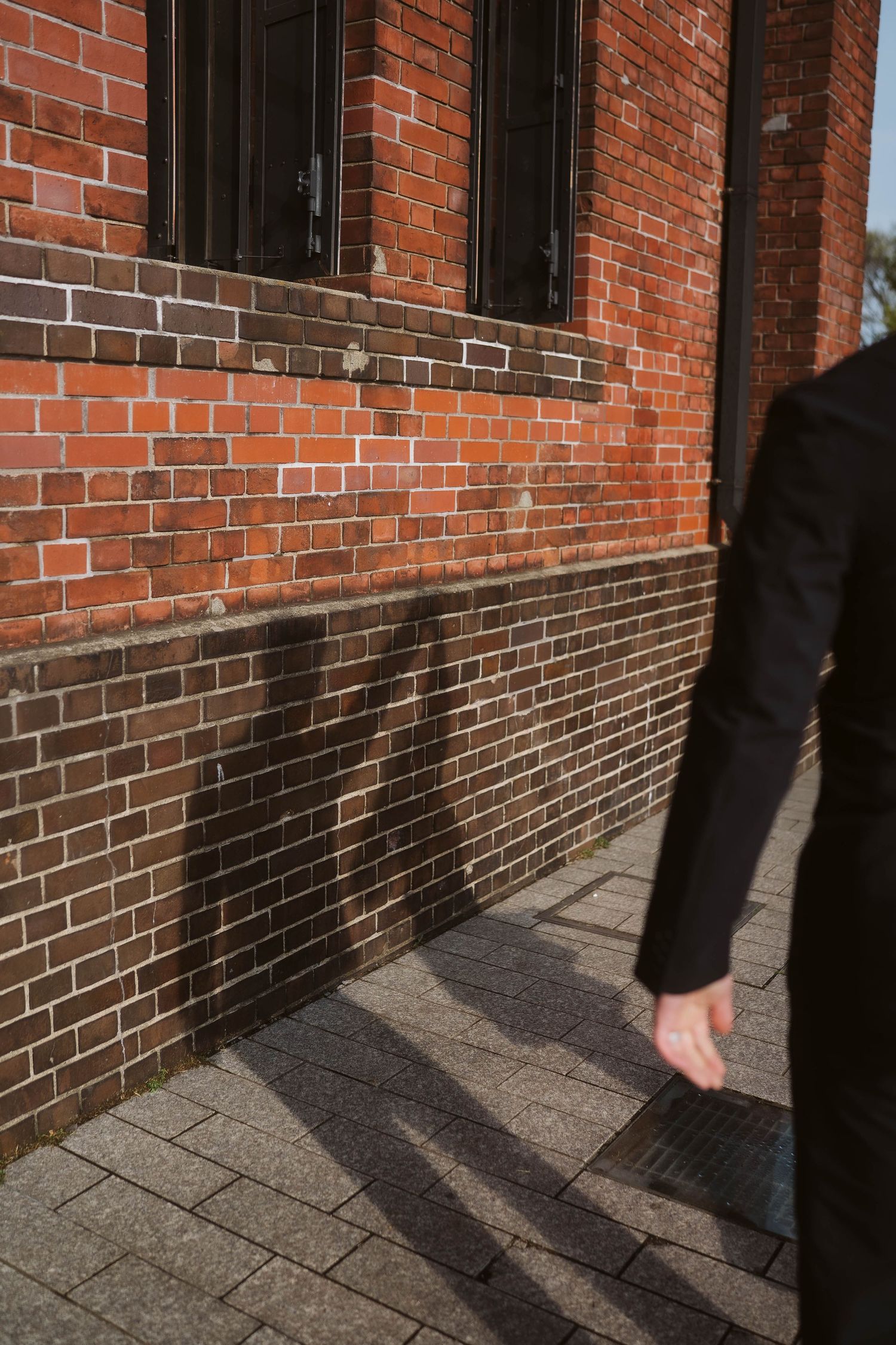 Partial view of a person walking past a red brick building with cast shadows on the pavement.