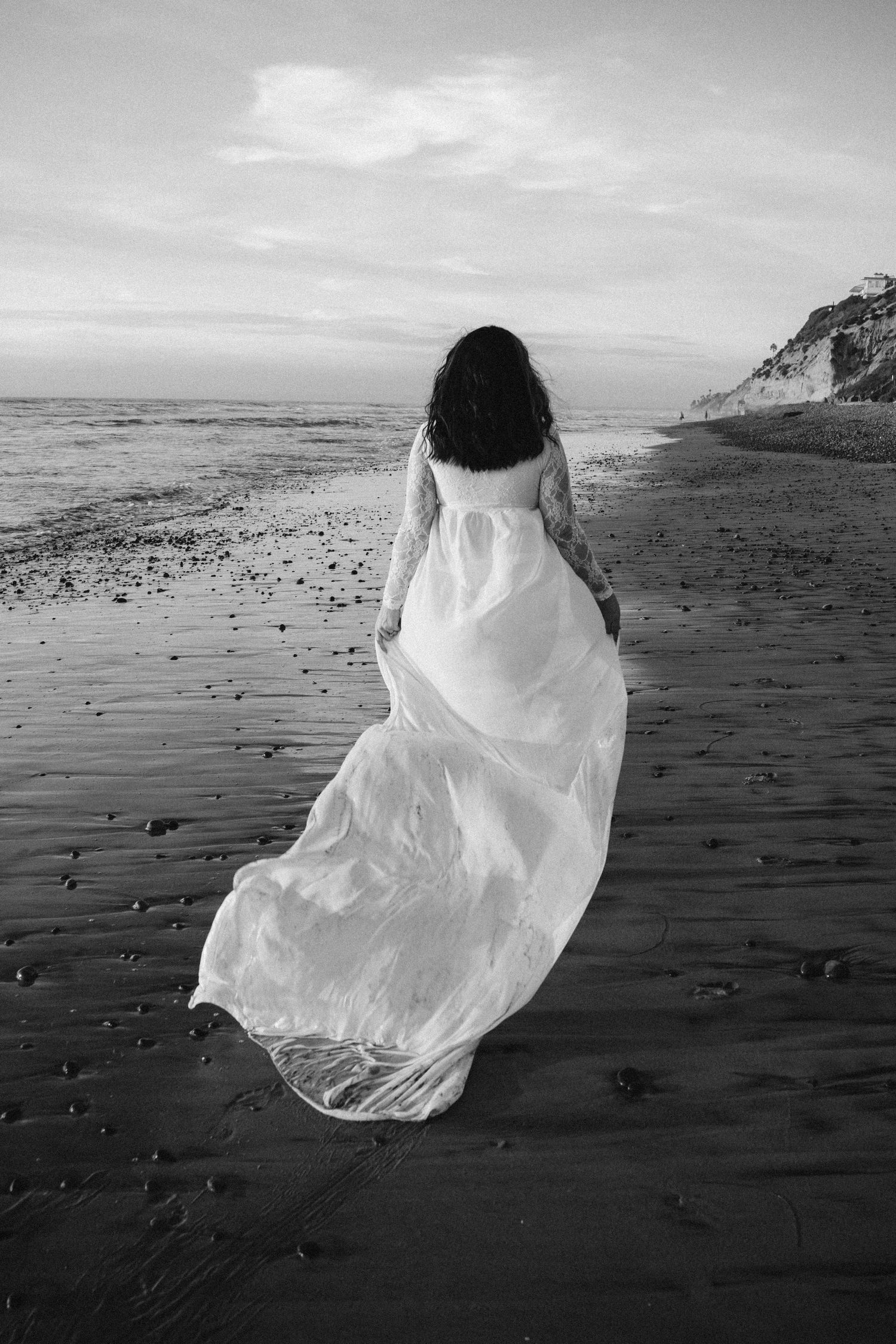 A figure in a flowing white dress walks along a misty beach shore with cliffs in the background.