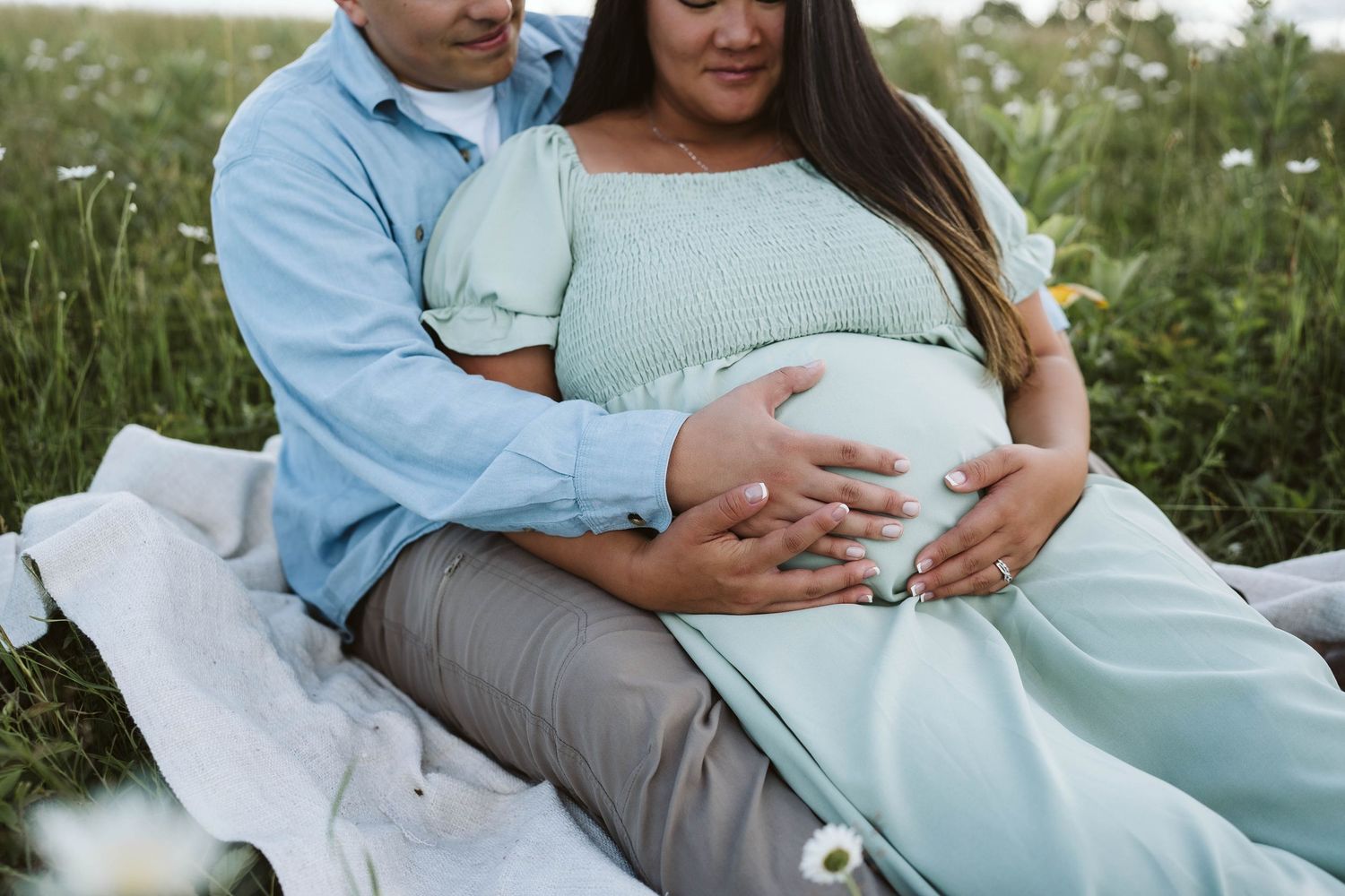 A pregnant couple sits on a blanket in a meadow of wildflowers wearing soft blue and gray colors.