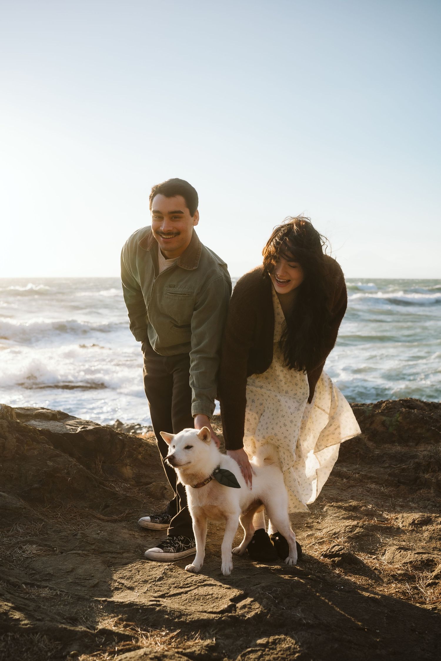 A couple and their white cat explore a rocky beach at sunset with ocean waves crashing in the background.