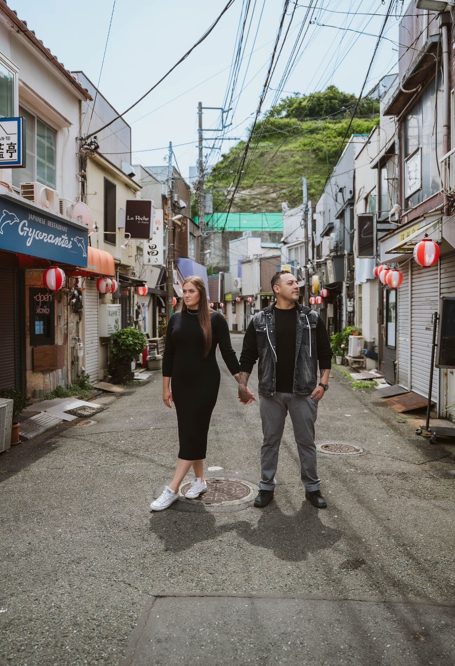 A couple explores a narrow Japanese street lined with traditional buildings and power lines.