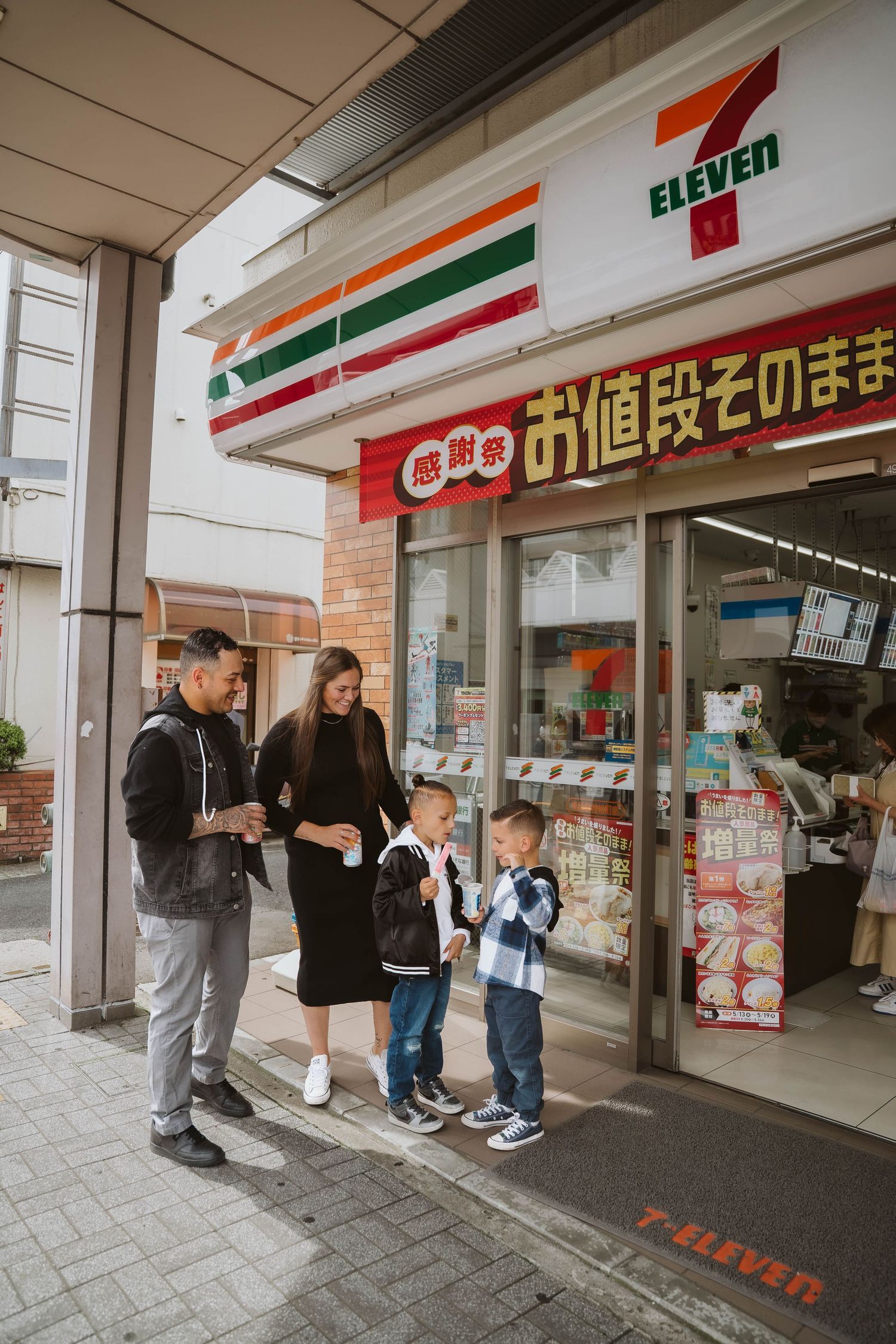 Group of people gathered outside a 7-Eleven storefront with Japanese signage in an urban setting.