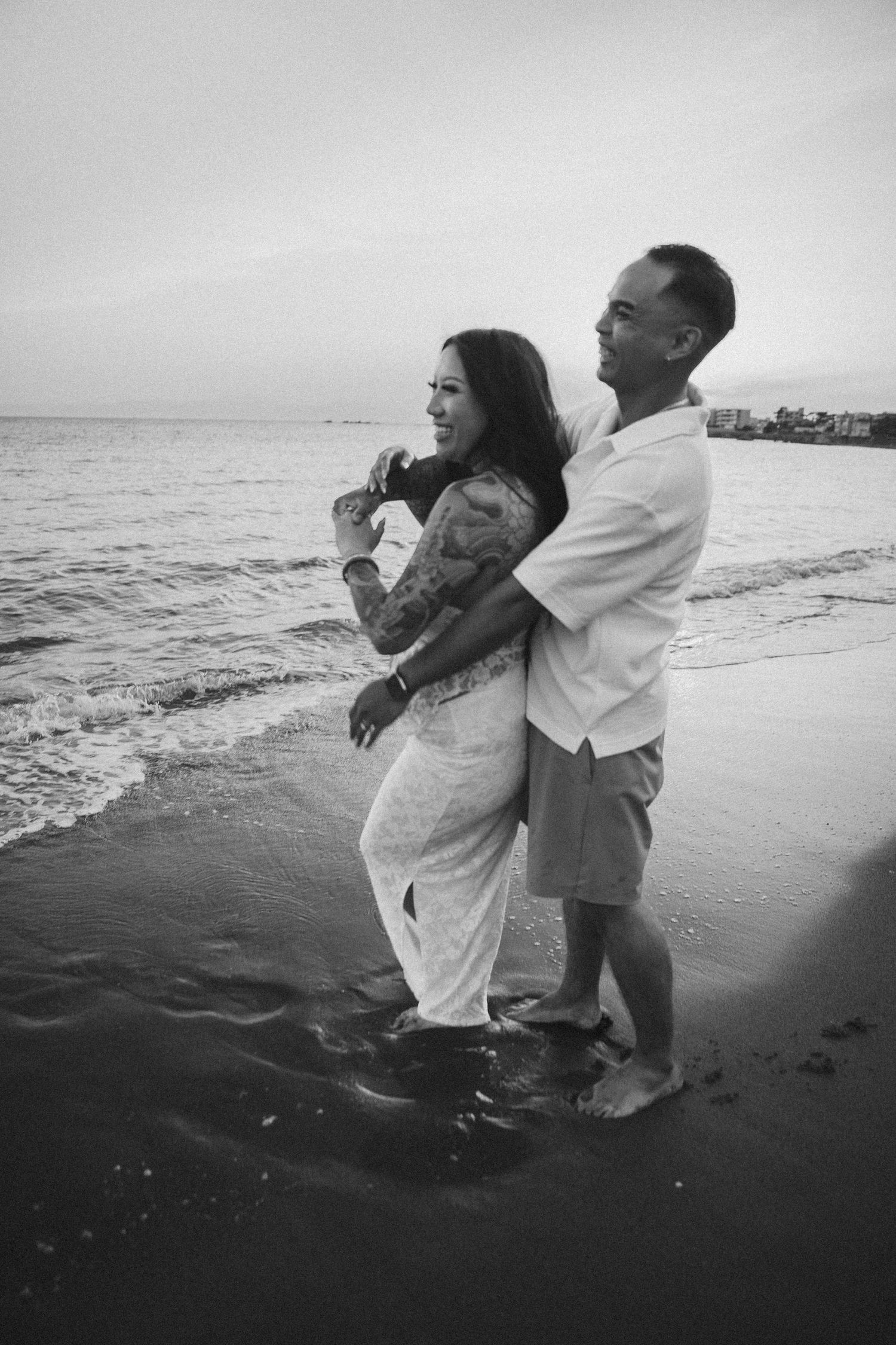 Black and white photo of a couple embracing on a beach with ocean waves in the background.