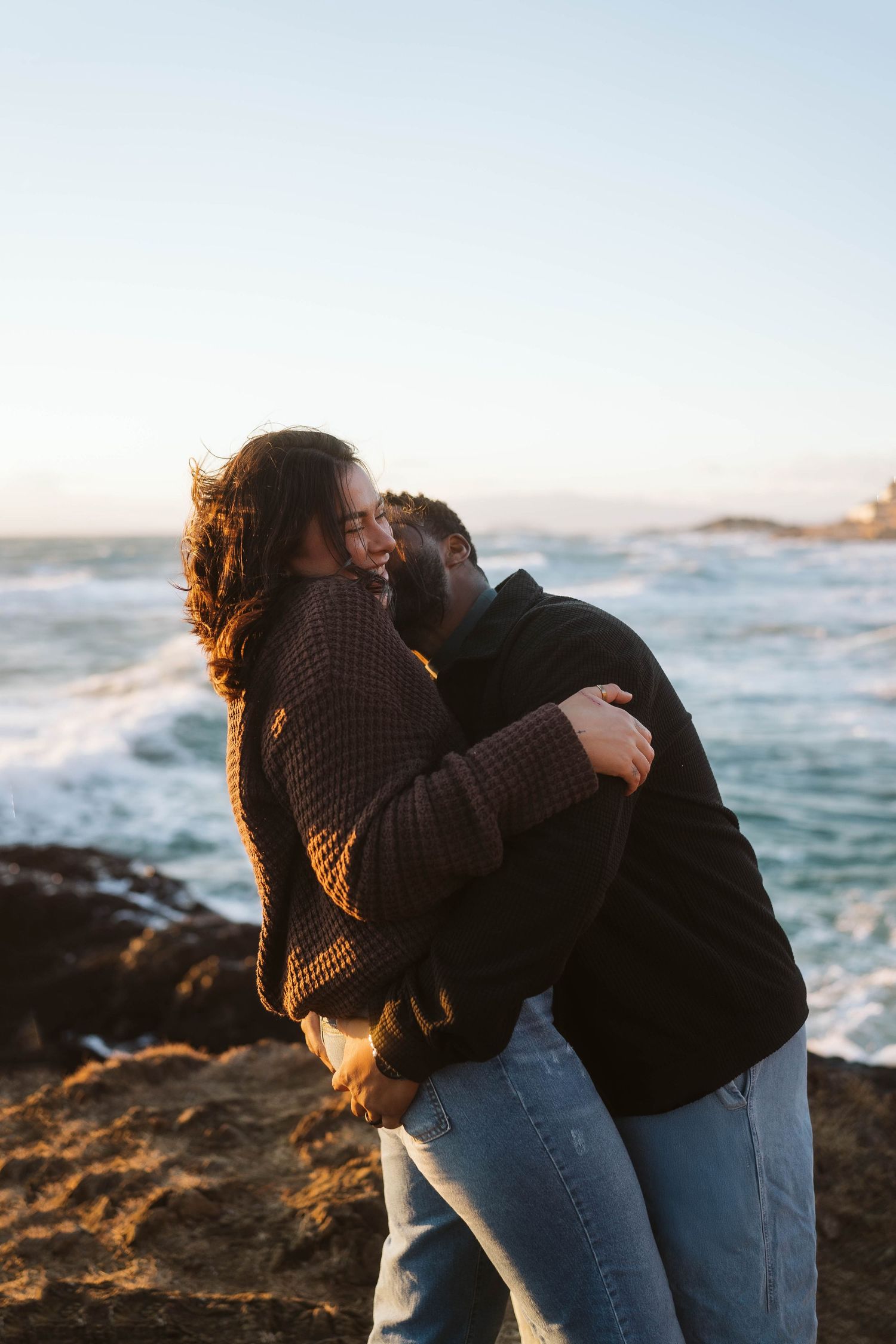 Two people embrace on a rocky coastline at sunset with ocean waves crashing in the background.