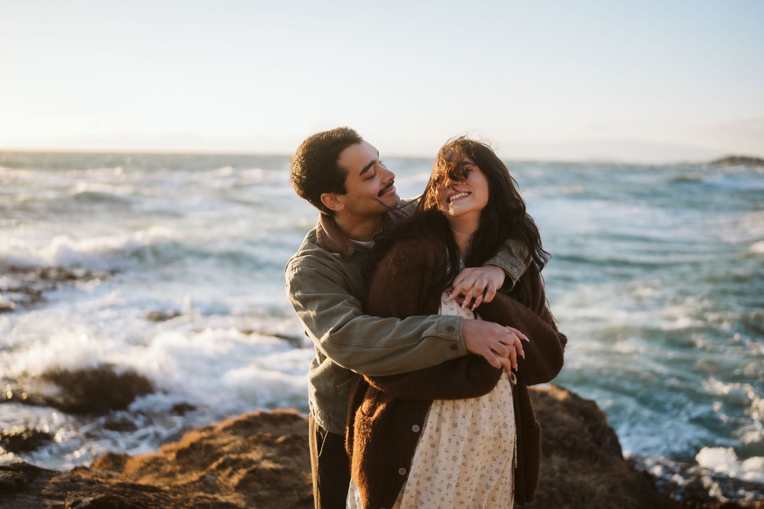 A romantic moment shared between a couple on rocky cliffs by the ocean during golden hour.