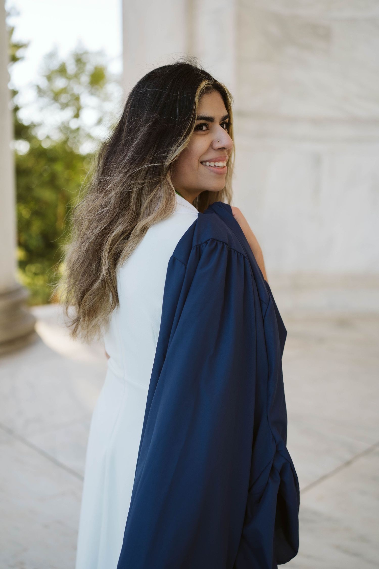 A graduate in navy blue cap and gown with long ombre hair smiles while looking over their shoulder during a photoshoot.