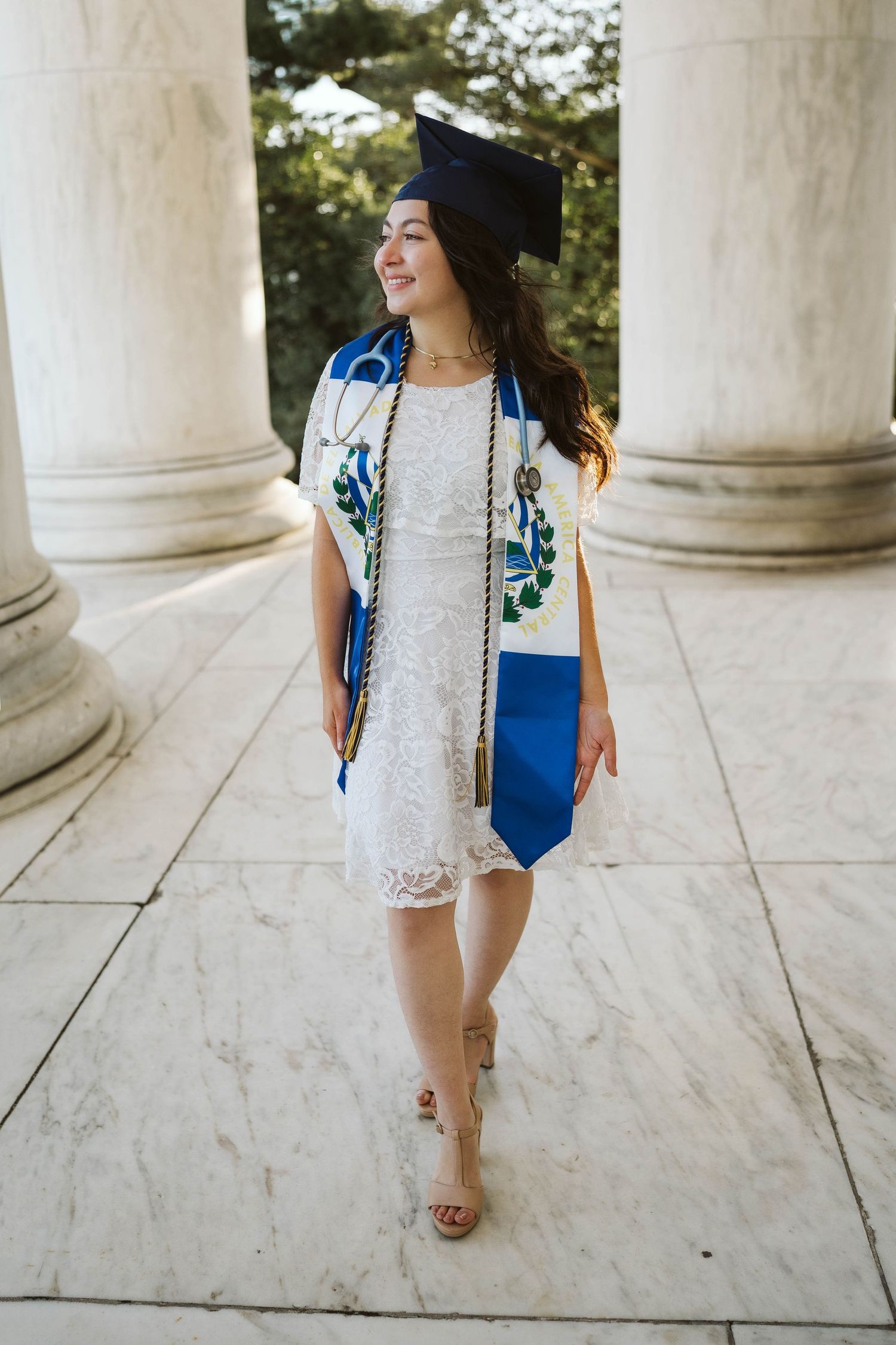 Graduate in white lace dress and blue stole walks between marble columns on graduation day.