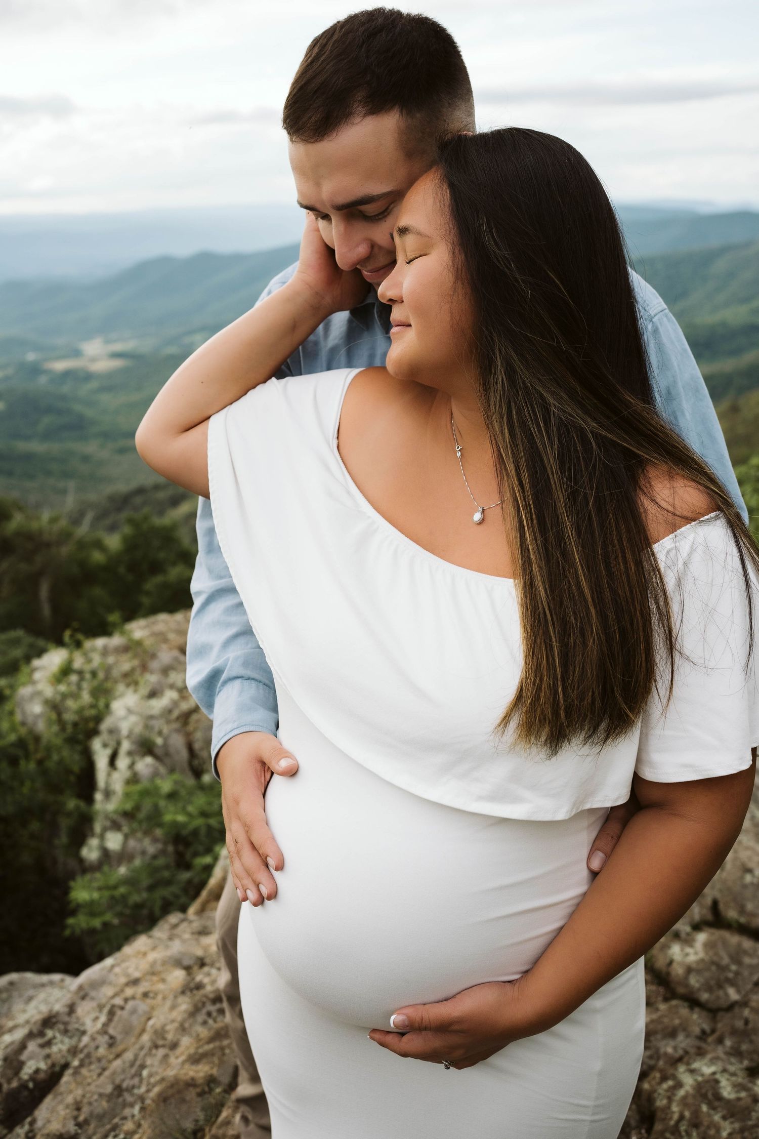 Couple embraces while standing on mountain overlook during maternity photo session in nature.
