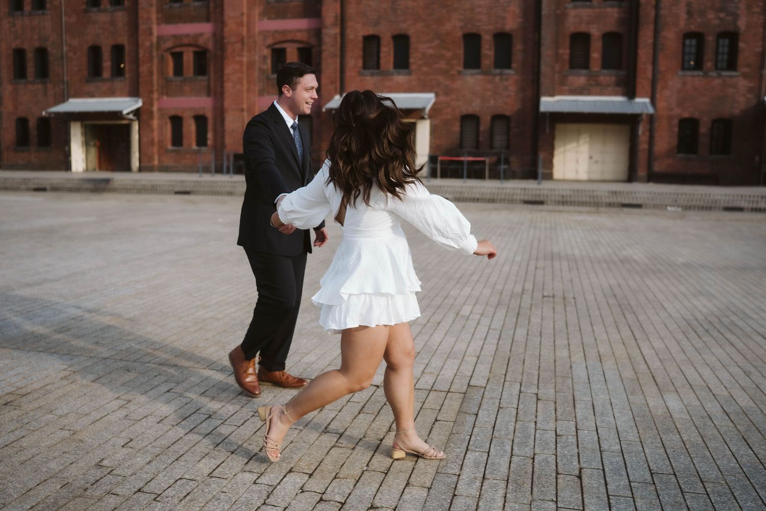 Couple dances joyfully on brick paved courtyard in formal attire with historic building facade in background.