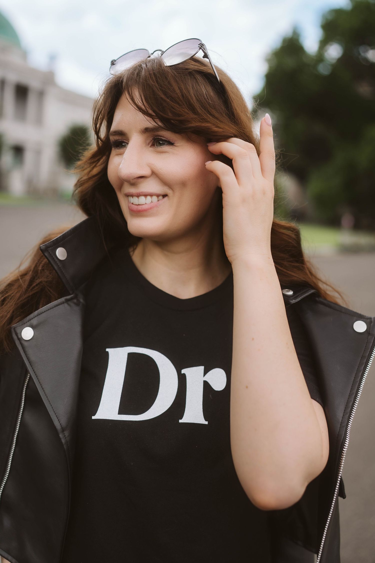 A person wearing a black leather jacket and sunglasses smiles while adjusting their glasses outdoors.
