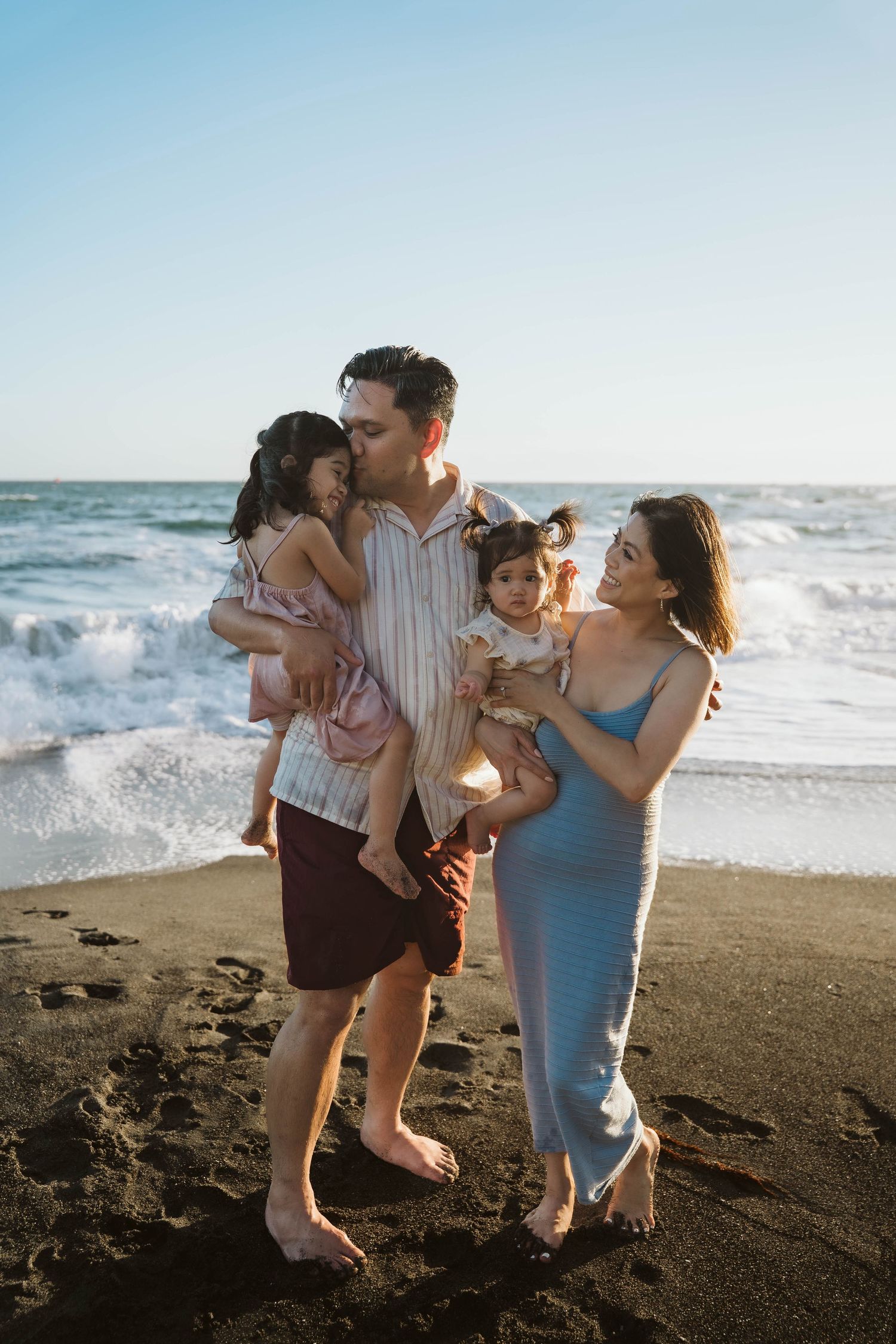 A loving family embraces while standing on a dark sandy beach during golden hour with waves behind them.