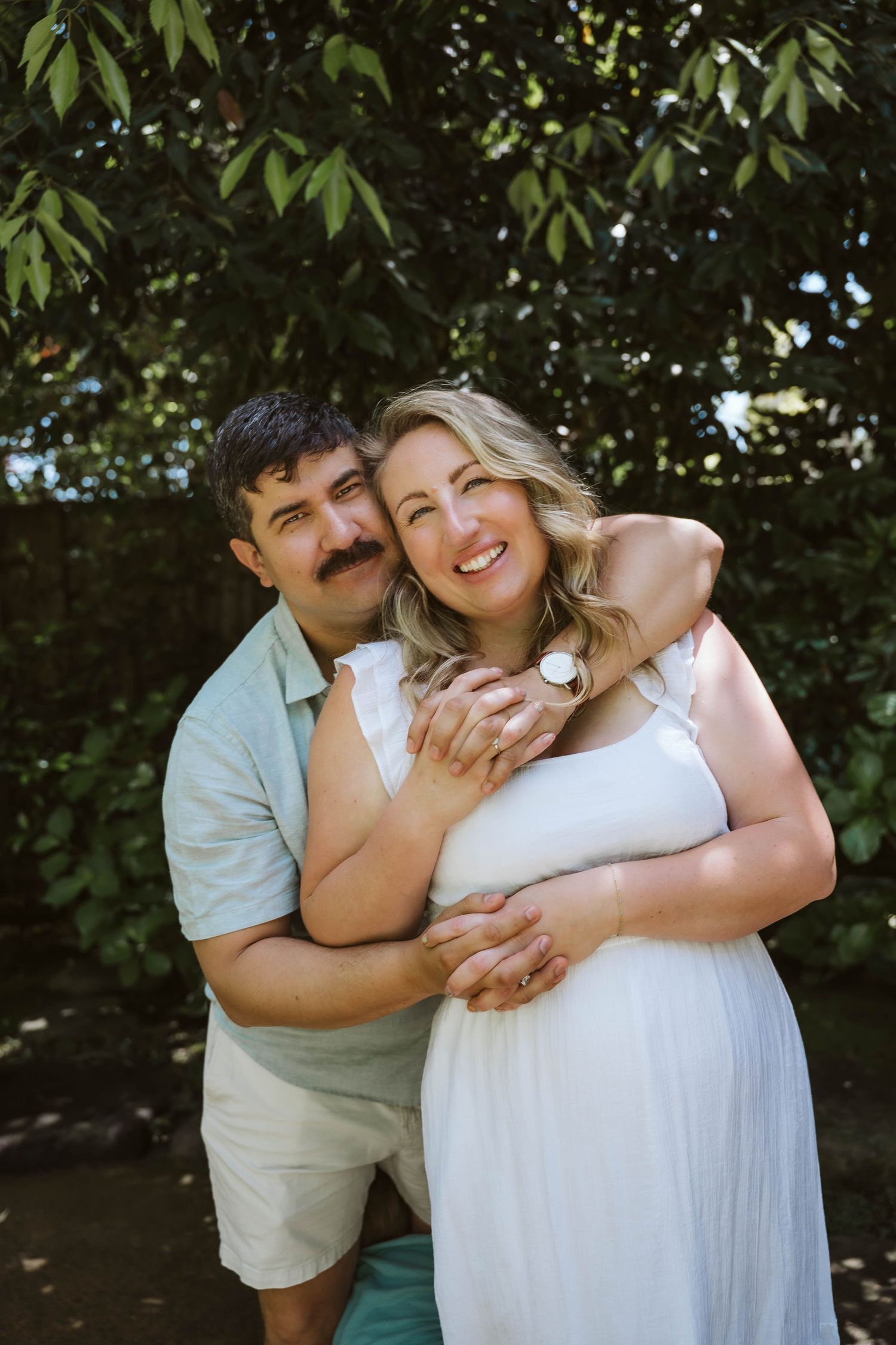 A couple embraces outdoors in a romantic photoshoot under green leafy trees.